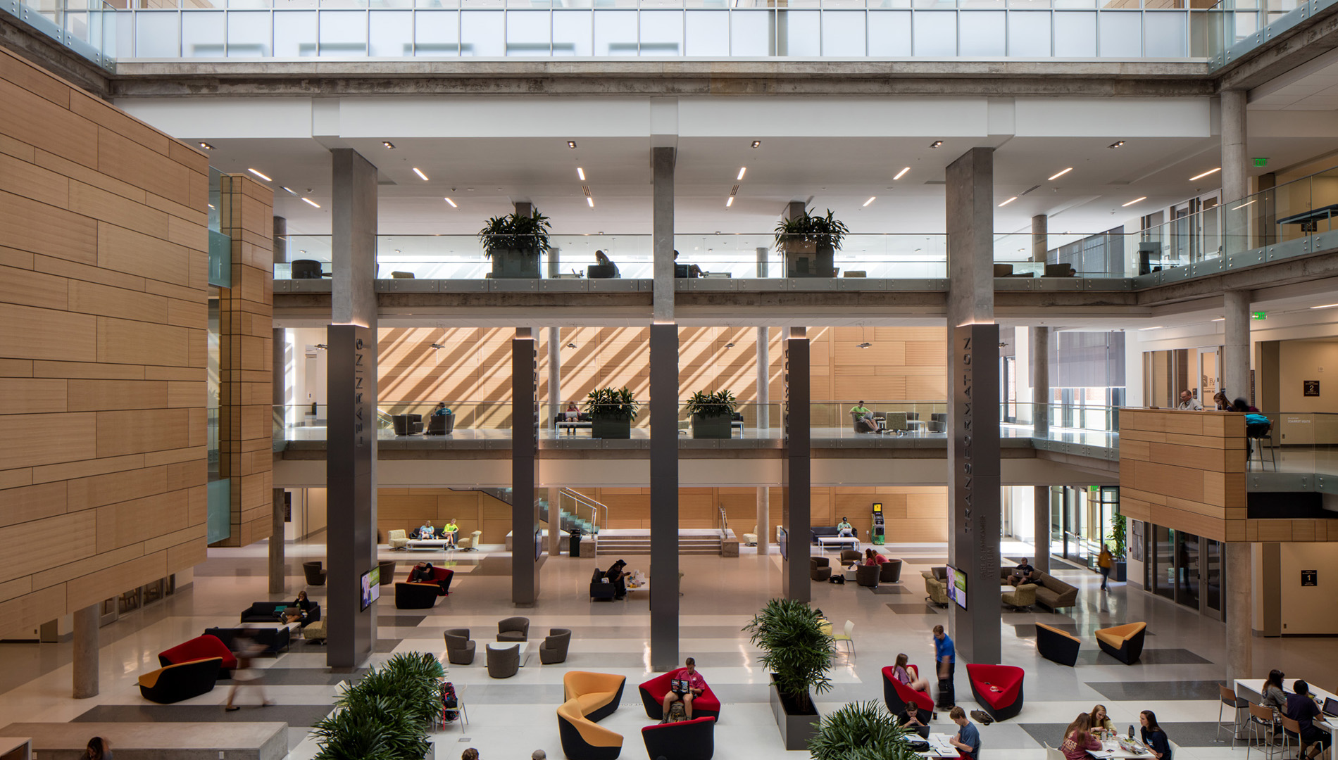 A spacious, modern atrium with multiple seating areas, columns, and two upper levels. People are working, conversing, and walking. Large windows admit natural light, and greenery decorates the space.