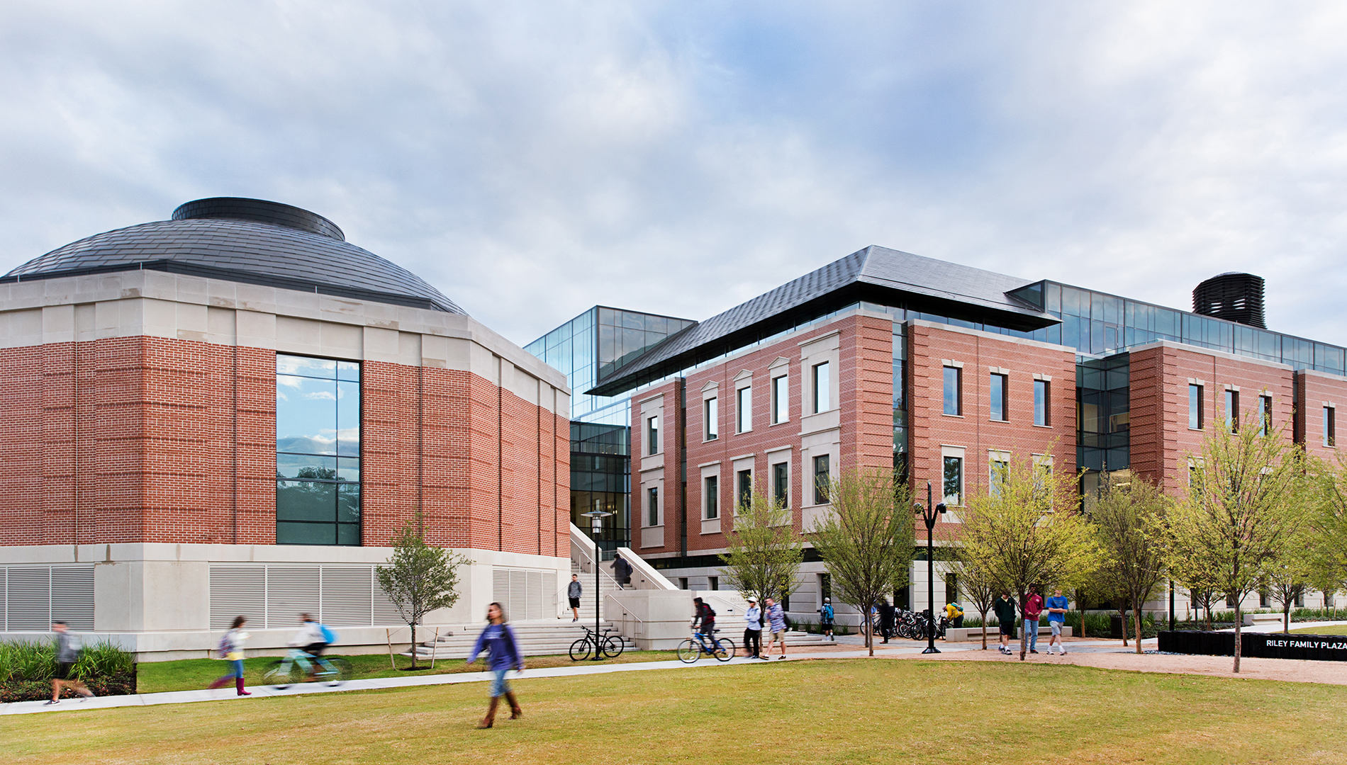 A modern academic building with a circular lecture hall (left) and a rectangular office and classroom wing (right). People walk and bike on the pathway in front. The sky is cloudy.