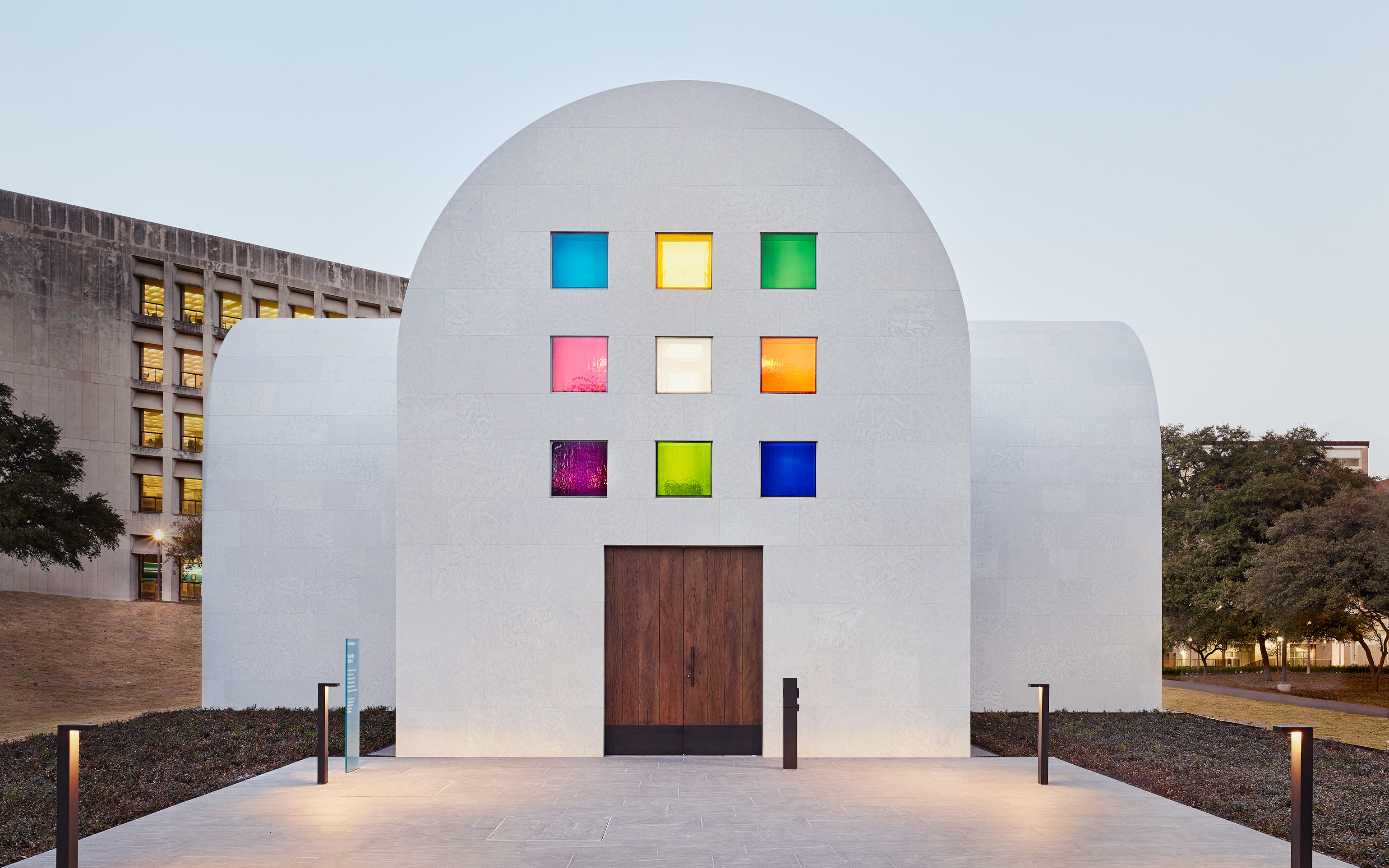 Modern building with a domed roof featuring a wooden door and a facade with nine square colorful windows. Adjacent are trees, another building, and a pathway leading to the entrance.