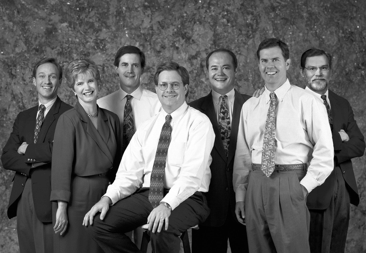 A group of six men and one woman in business attire pose for a professional group photo against a textured backdrop, with Mark Barnet Headley standing front and center.