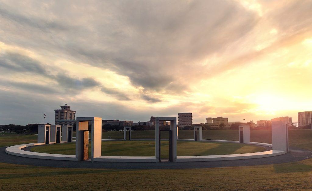 A circular arrangement of tall, rectangular stone pillars on a grassy area with a city skyline and a cloudy sky at sunset in the background resembles a serene bonfire memorial.