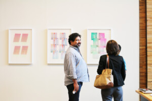 Two people stand in front of a wall adorned with three framed abstract artworks. One person faces the art while the other, wearing a gray shirt, looks toward the camera. The pieces are part of the Overland Marks art program featuring Texas artists.