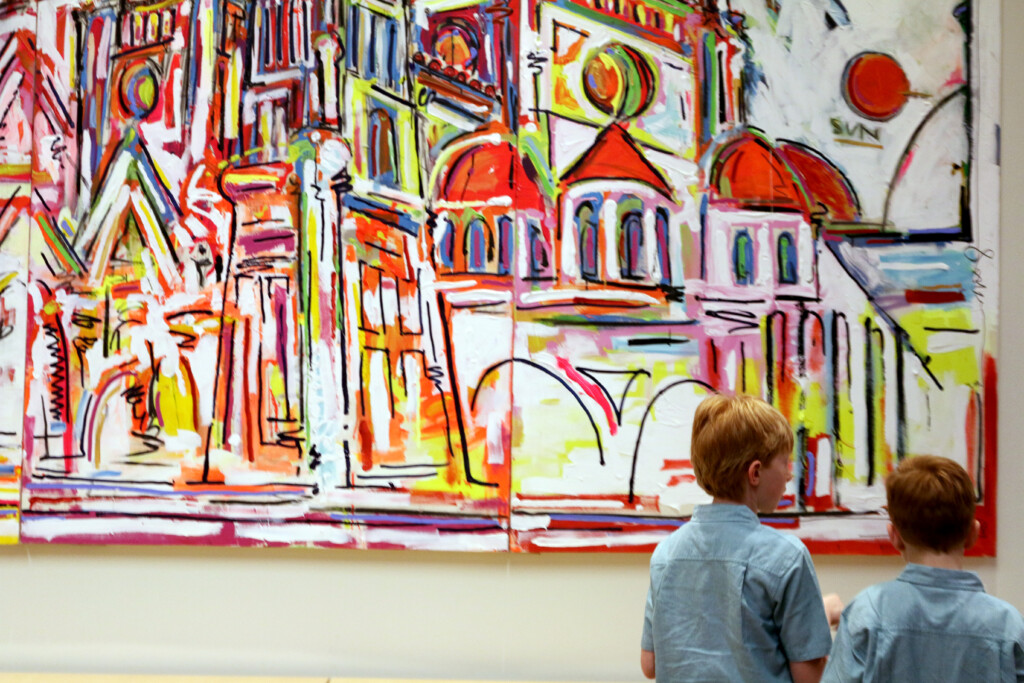 Two boys stand in front of a large, colorful abstract painting by Texas artists, featuring buildings and architectural elements with red domes.