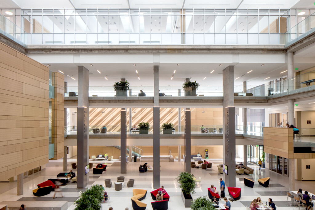 A spacious, modern atrium with multiple floors, glass walls, and various seating areas at Baylor University's Foster Campus. People are scattered around, engaged in different activities. Plants and decor add greenery and color to the resilient design.