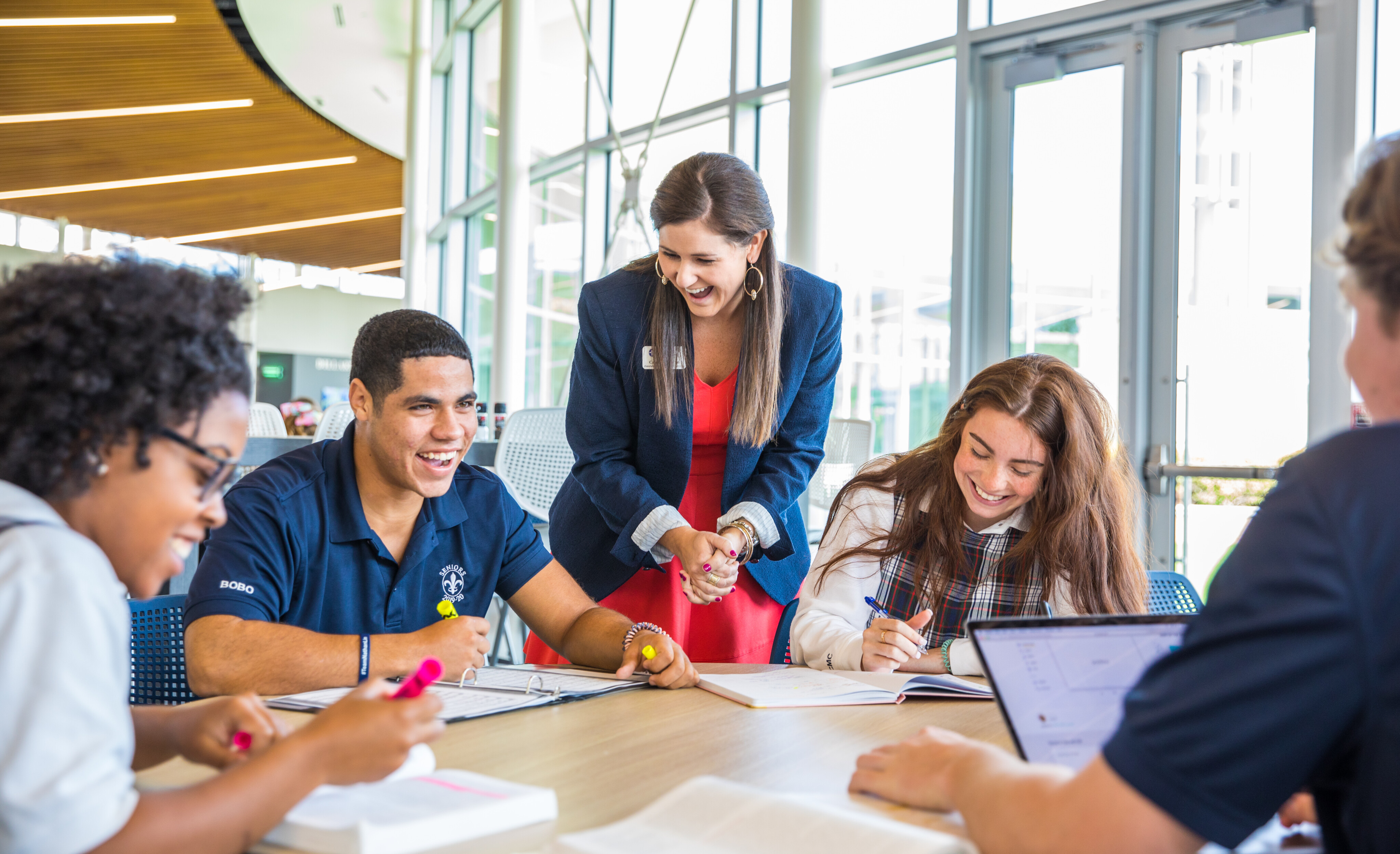 A group of students sits at a table working on papers while a teacher, standing, engages with them in a brightly lit physical space.