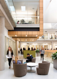 Modern, multi-level office space with people working, walking, and interacting; features glass railings, wooden accents, and indoor plants. Three people sit in gray chairs around a circular table in a setting reminiscent of the resilient design seen at Baylor University's Foster Campus.