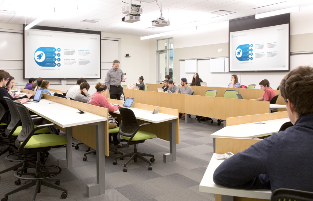 A classroom at Baylor University with students seated at desks, listening to an instructor. The instructor stands at the front near a projector screen displaying a presentation on 21st Century Learning. Several students have laptops open, integrating modern technology into their resilient design of education.