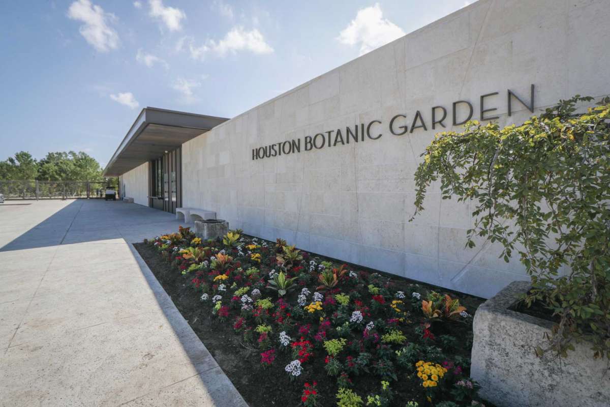 The entrance of the Houston Botanic Garden, designed by Overland Partners, boasts beige stone walls, large windows, and an adjacent garden bed filled with colorful flowers under a clear blue sky.