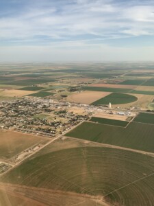 Aerial view of a rural landscape featuring agricultural fields, a small town reminiscent of Downtown Lubbock, and circular crop patterns under a partly cloudy sky.