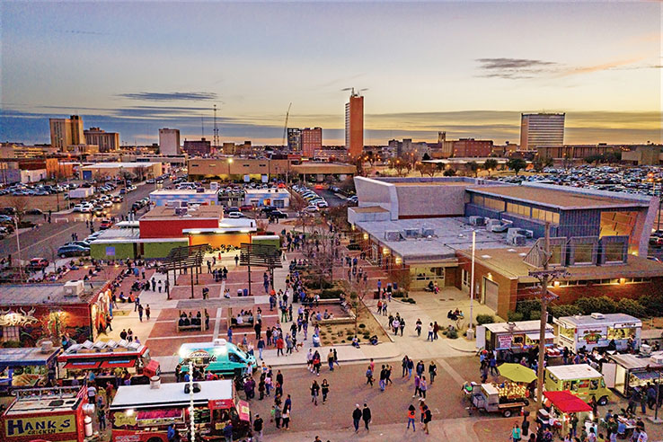 Aerial view of a bustling urban area at sunset in Downtown Lubbock, with food trucks, a large crowd, multiple buildings, and the city skyline in the background.