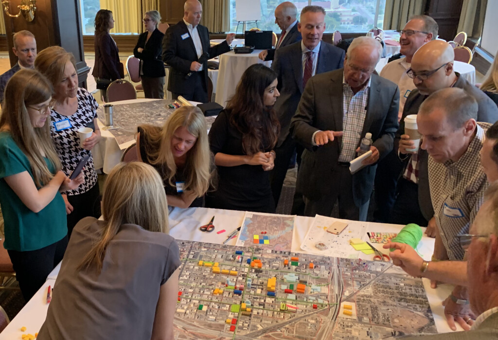 A group of people are gathered around a table looking at a large map with various colored blocks. Some are standing, others are seated, and all appear engaged in discussion about catalyzing the transformation of downtown Lubbock.