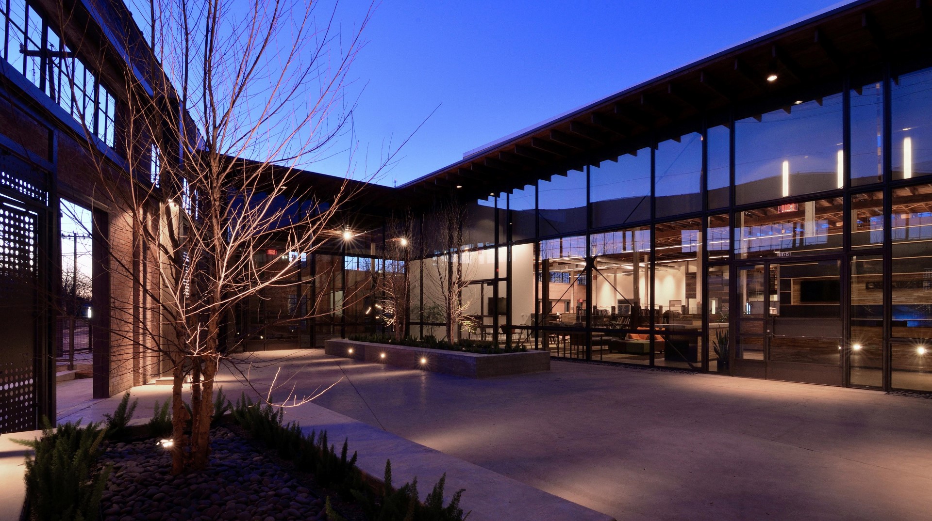 Modern architecture by Overland Partners featuring an open courtyard with planters, bare trees, and a building with large glass windows showcasing an illuminated interior at dusk.