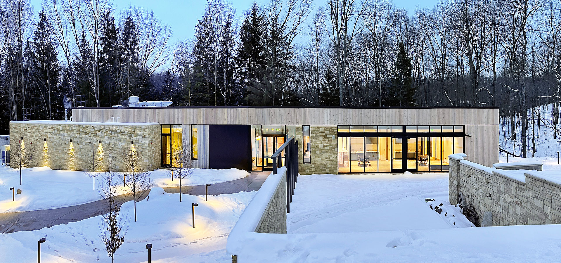 A modern building stands amidst a snowy landscape, illuminated with warm yellow lights. Leafless trees and nightfall add a serene winter ambiance.