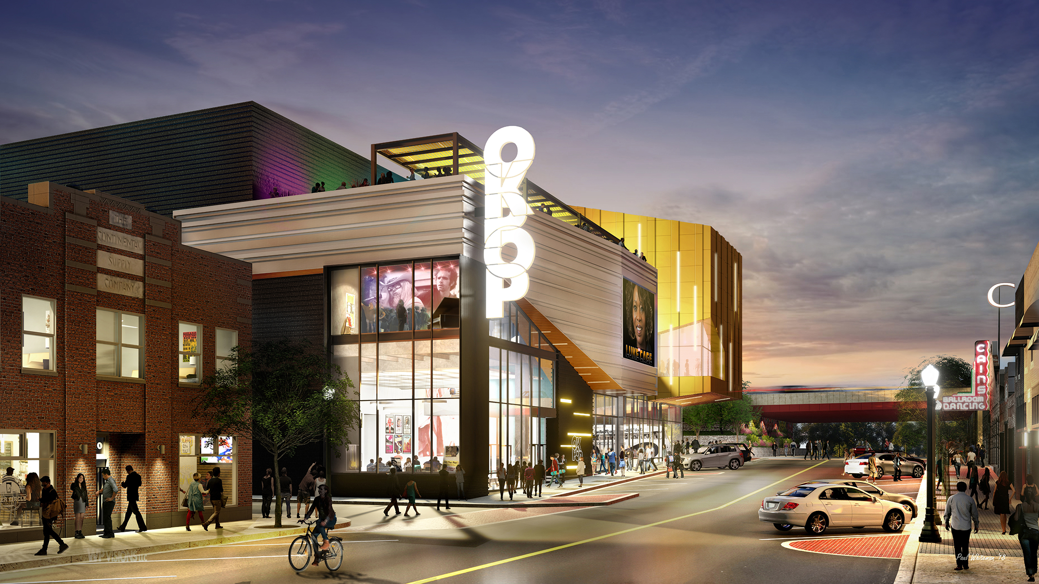 Street view of a modern illuminated building with an "OSU" sign, showcasing a mix of retail and entertainment spaces. People walk and cycle on the streets, and cars are parked along the roadside. The vibrant scene is reminiscent of the nearby Oklahoma Museum of Popular Culture (OKPOP).