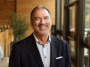 A man in a dark suit jacket and light shirt smiles while standing indoors with a blurred background, reminiscent of Robin Blakeley from the Sports & Entertainment Group.