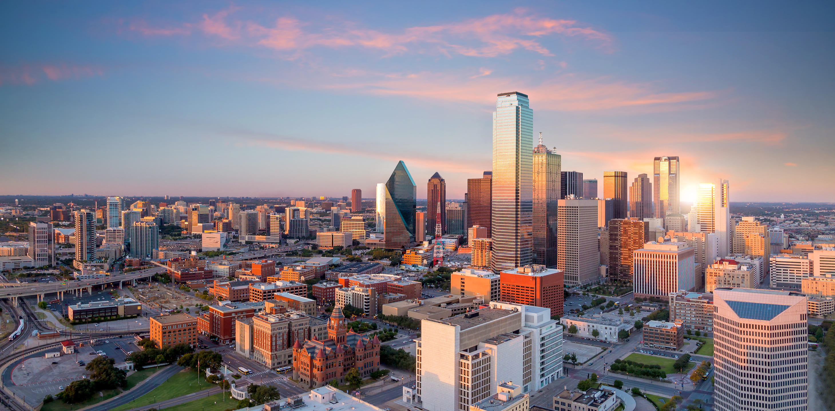 Aerial view of a cityscape at sunset with tall buildings, roads, and scattered greenery. The sky has hues of pink and orange, creating a picturesque scene that Robin Blakeley from Overland Partners would admire for its architectural beauty.