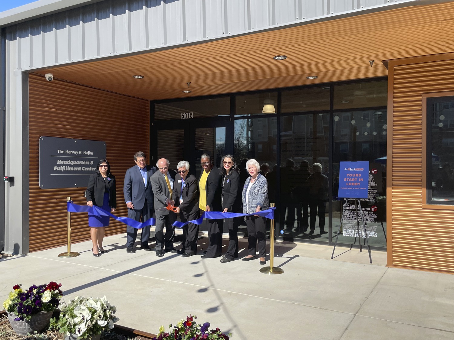 A group of people stands behind a blue ribbon in front of a building entrance, preparing for a grand opening ribbon-cutting ceremony. A sign nearby reads, "Headquarters & Fulfillment Center," highlighting the collaboration with Overland Partners and Project Mend.