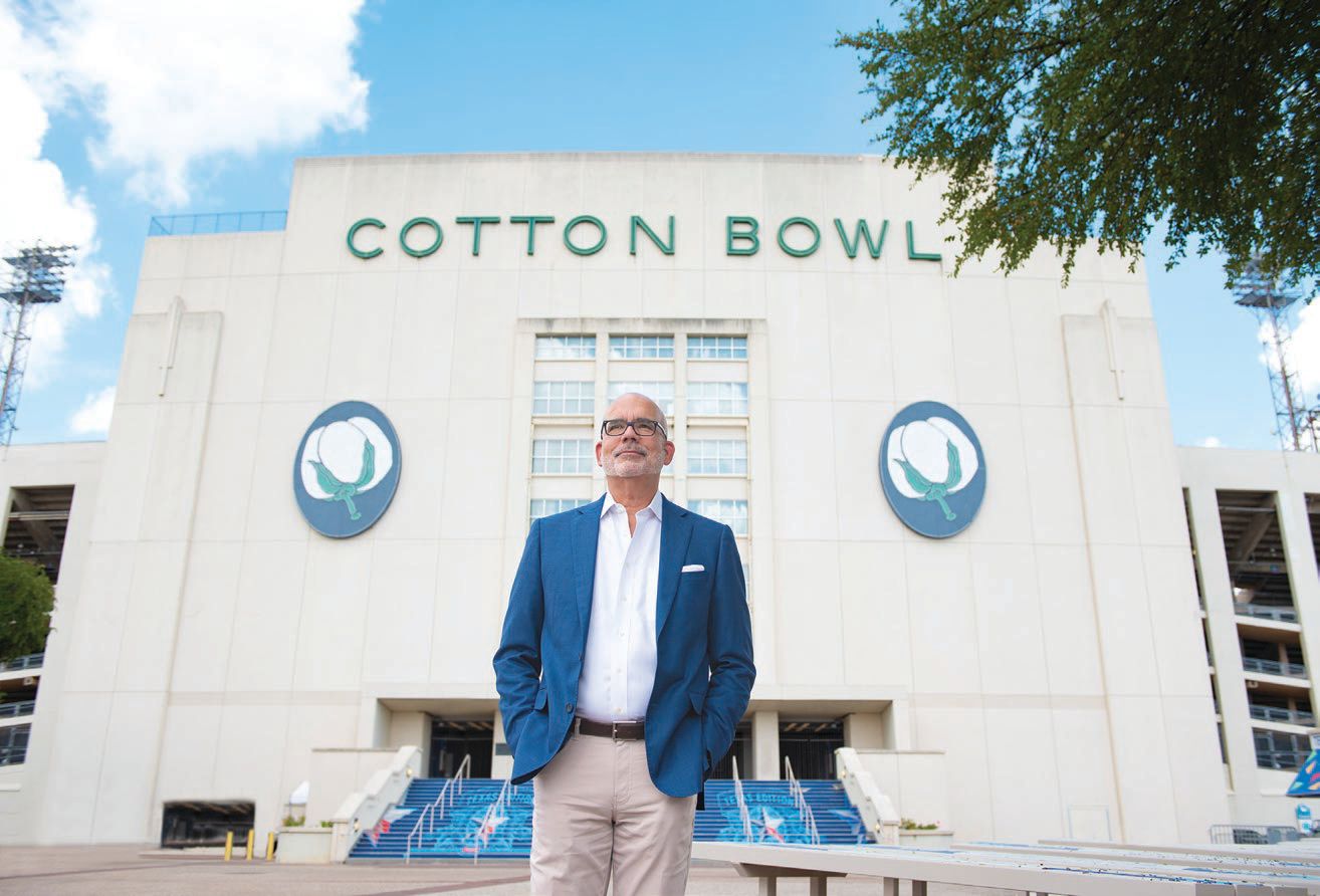 A man in a blue blazer and white shirt stands in front of the Cotton Bowl stadium, designed by Bryan Trubey, with its name and cotton logos visible on the exterior wall.