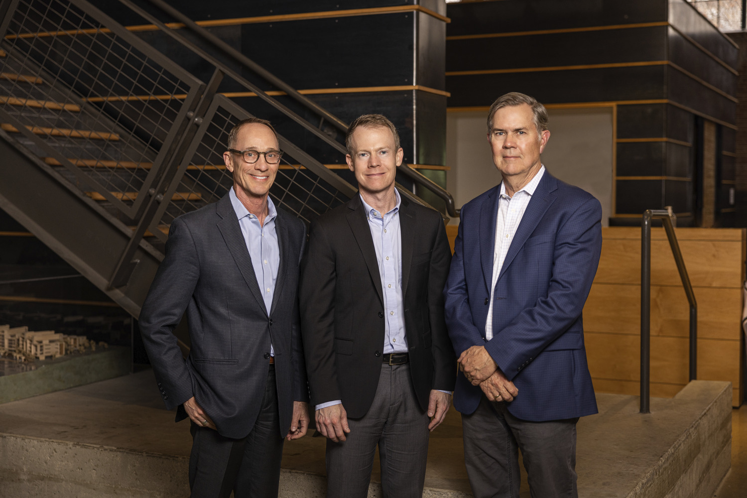 Three men in business attire stand side by side in a modern office setting, representing the Overland leadership team. The space features a staircase and wooden accents, reflecting the company's innovative spirit.