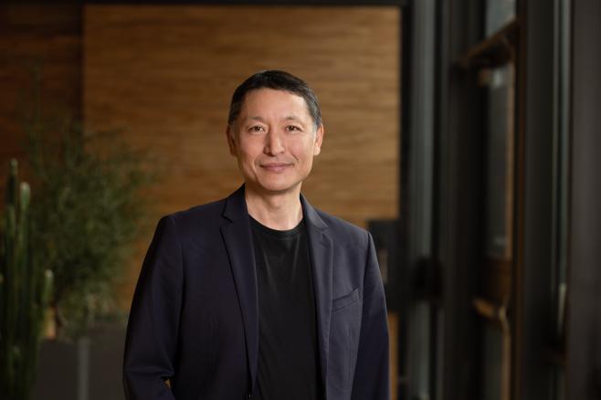 A person wearing a dark blazer and black shirt stands indoors with a neutral expression. The background shows a wooden wall and large windows.