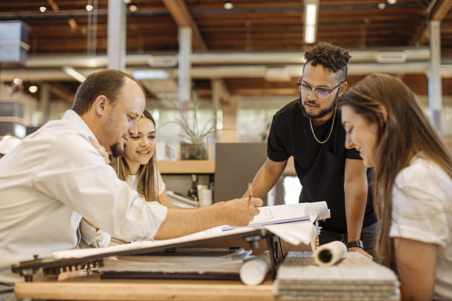 Four people collaborate around a desk with documents and blueprints in an open office environment, discussing or reviewing the materials. Their transparent communication fuels innovative ideas for Overland projects, fostering a brighter future for everyone involved.