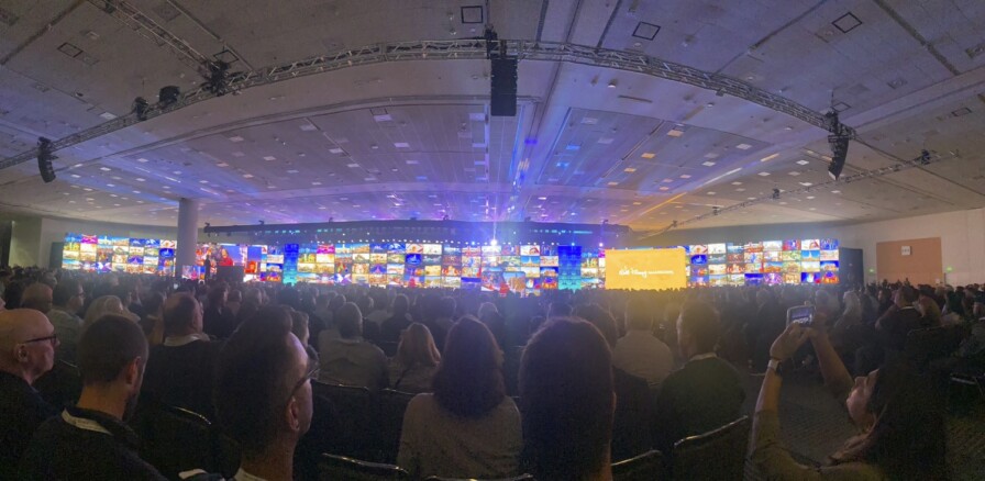 A large crowd sits in an auditorium filled with colorful screens and a prominent stage displaying insights from the AIA National Convention 2023.