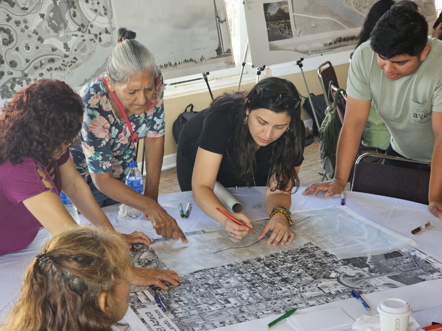 A group of people, including an elderly woman and a young man, gather around a table looking at a large map. One woman in black, from the UTSA Graduate Studio, is writing on the map while others attentively observe this overland expedition planning session.