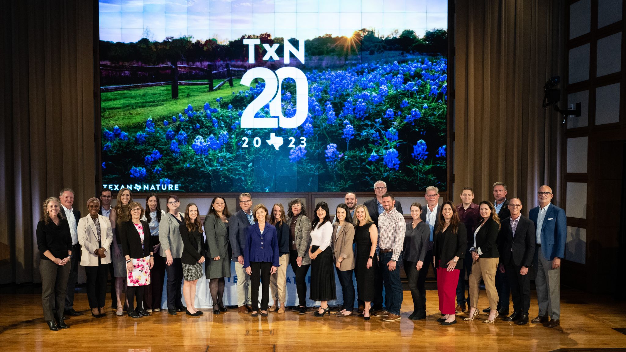 A group of 26 people stands on a wooden stage in front of a large screen displaying "Texan by Nature 20 2023" and an image of a field of bluebonnets.