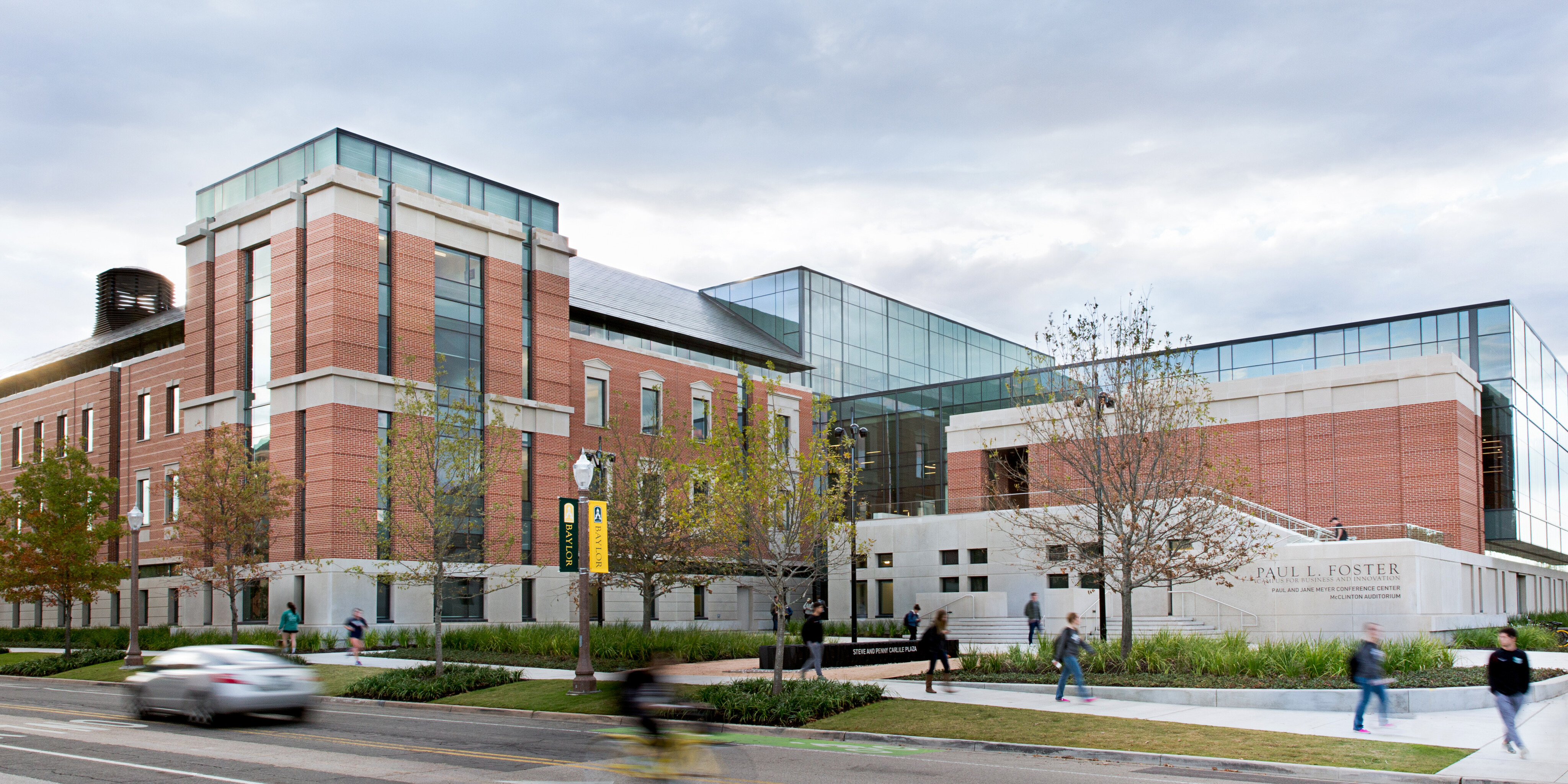 A modern brick and glass building houses Baylor's School of Business with pedestrians and cyclists passing by on the street in front. The sky is cloudy, and some greenery surrounds the building.