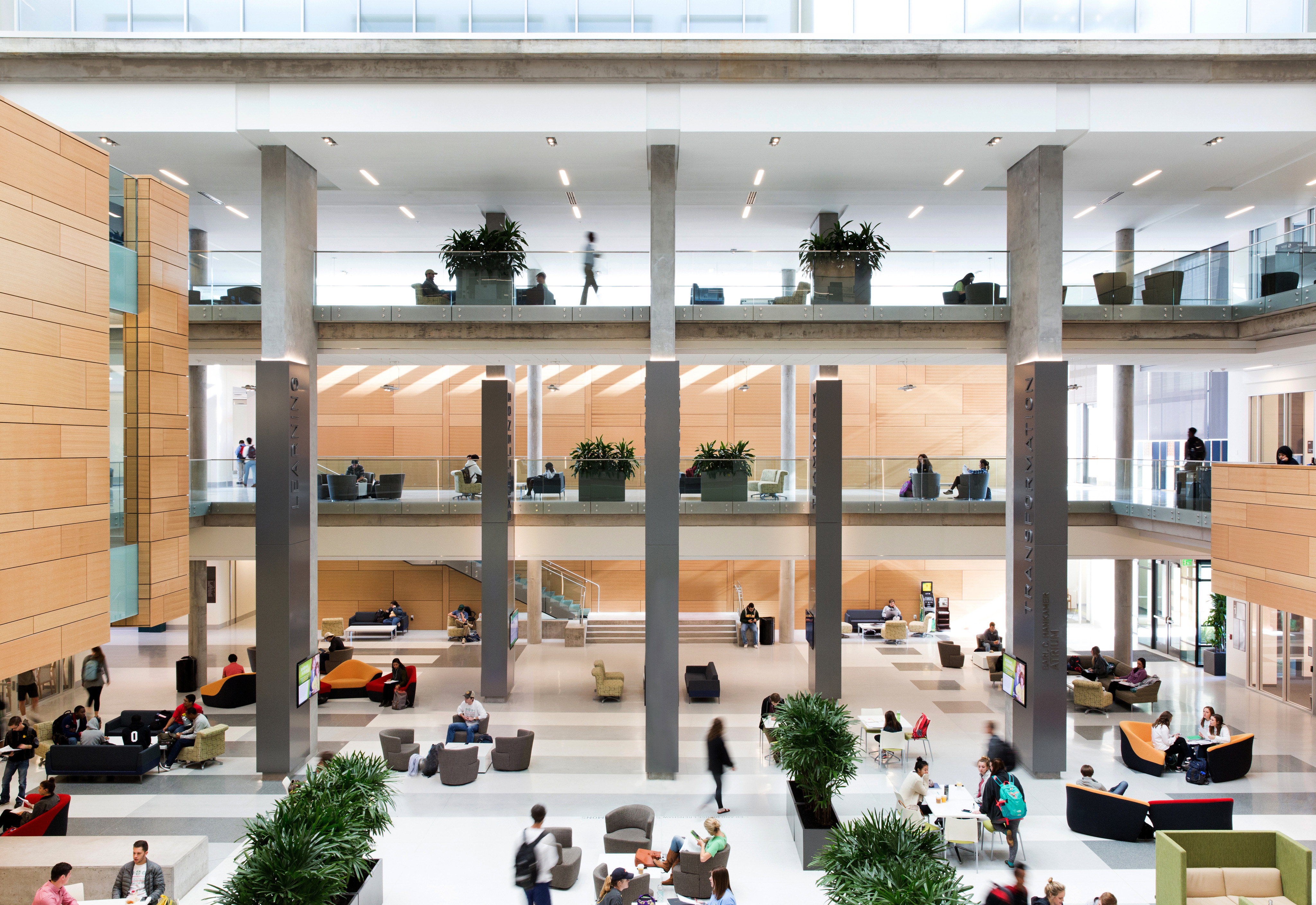 A modern, multi-level indoor public space with glass railings, tall columns, and people seated on various chairs and sofas. Natural light fills the atrium of Baylor's School of Business, with several plants placed throughout the area.