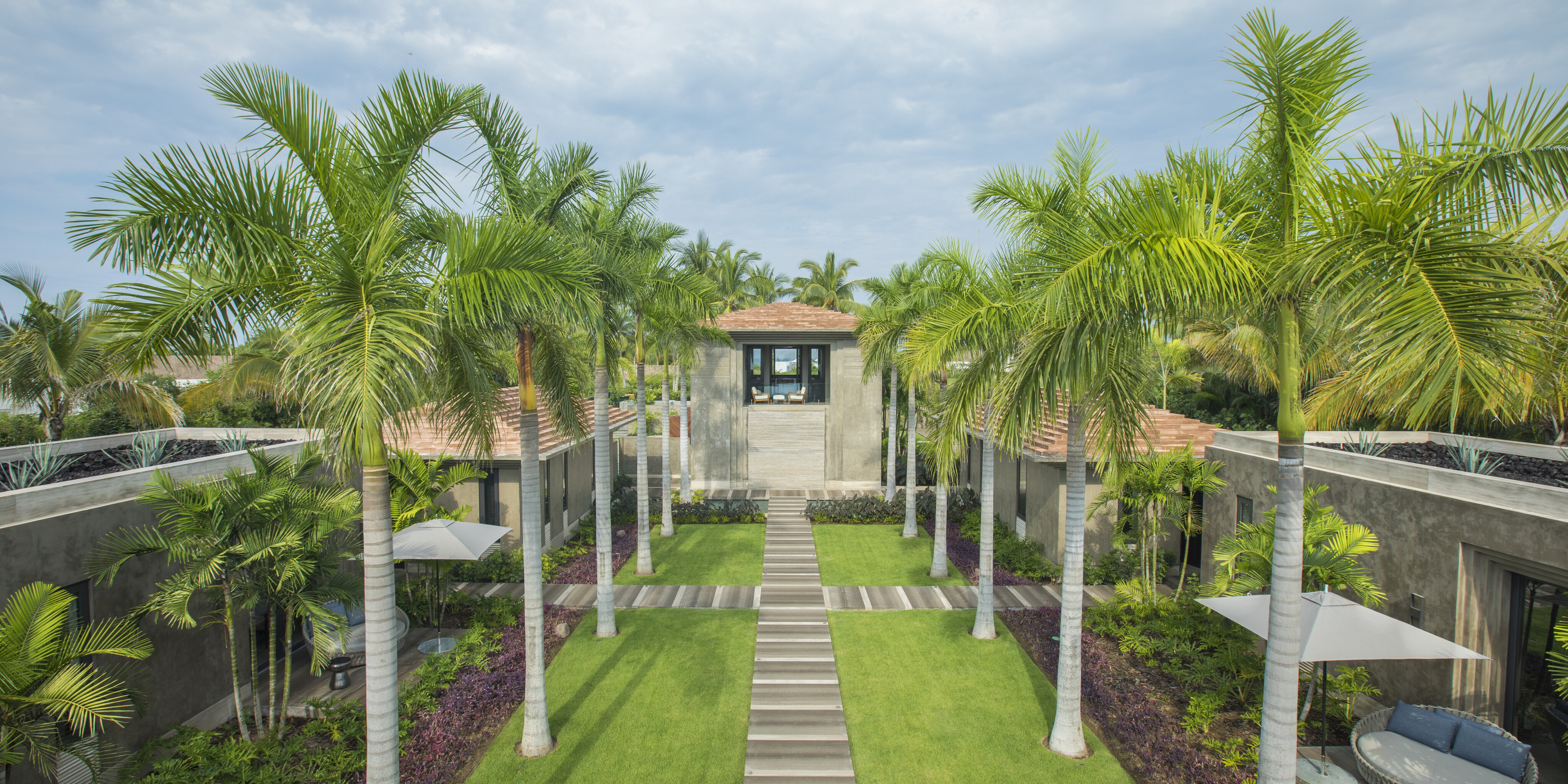 A well-maintained garden with a central pathway, surrounded by palm trees and greenery, leads to La Cascada, a restaurant with large windows under a cloudy sky.