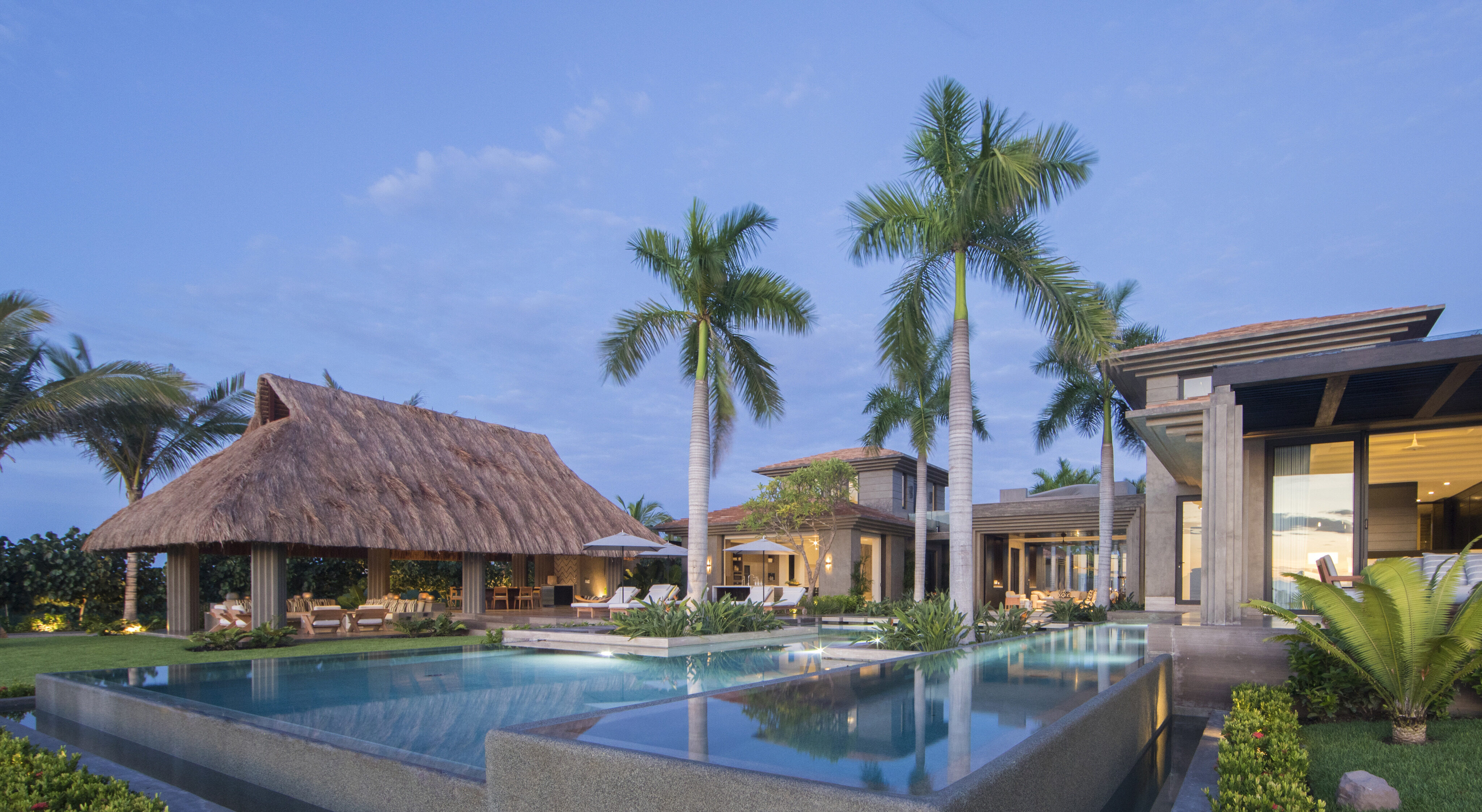 A luxury tropical villa, La Cascada, with a thatched roof cabana, palm trees, and an infinity pool at dusk.