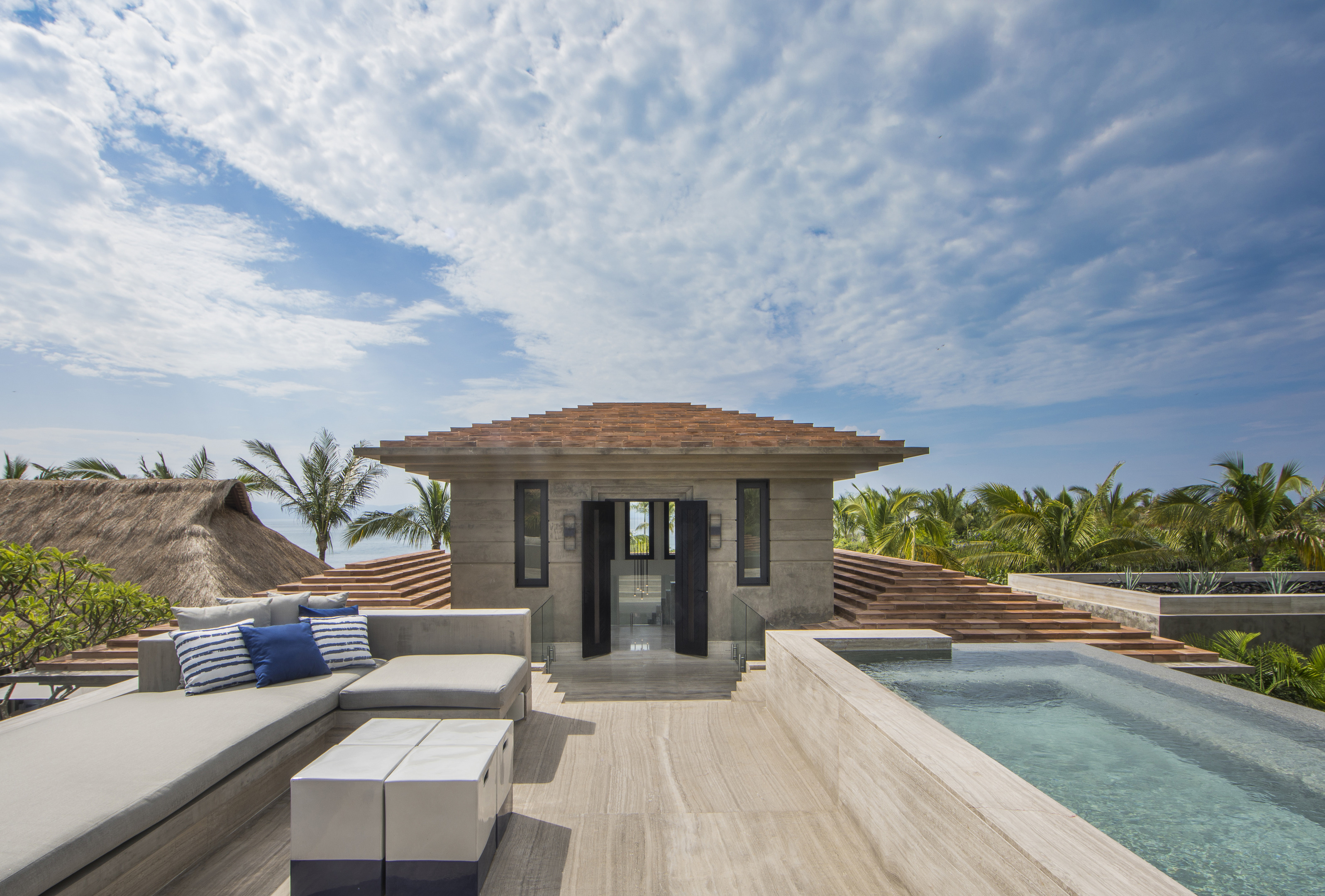 A modern terrace at La Cascada features a cozy seating area with cushions and a small pool, surrounded by tropical vegetation under a partly cloudy sky.