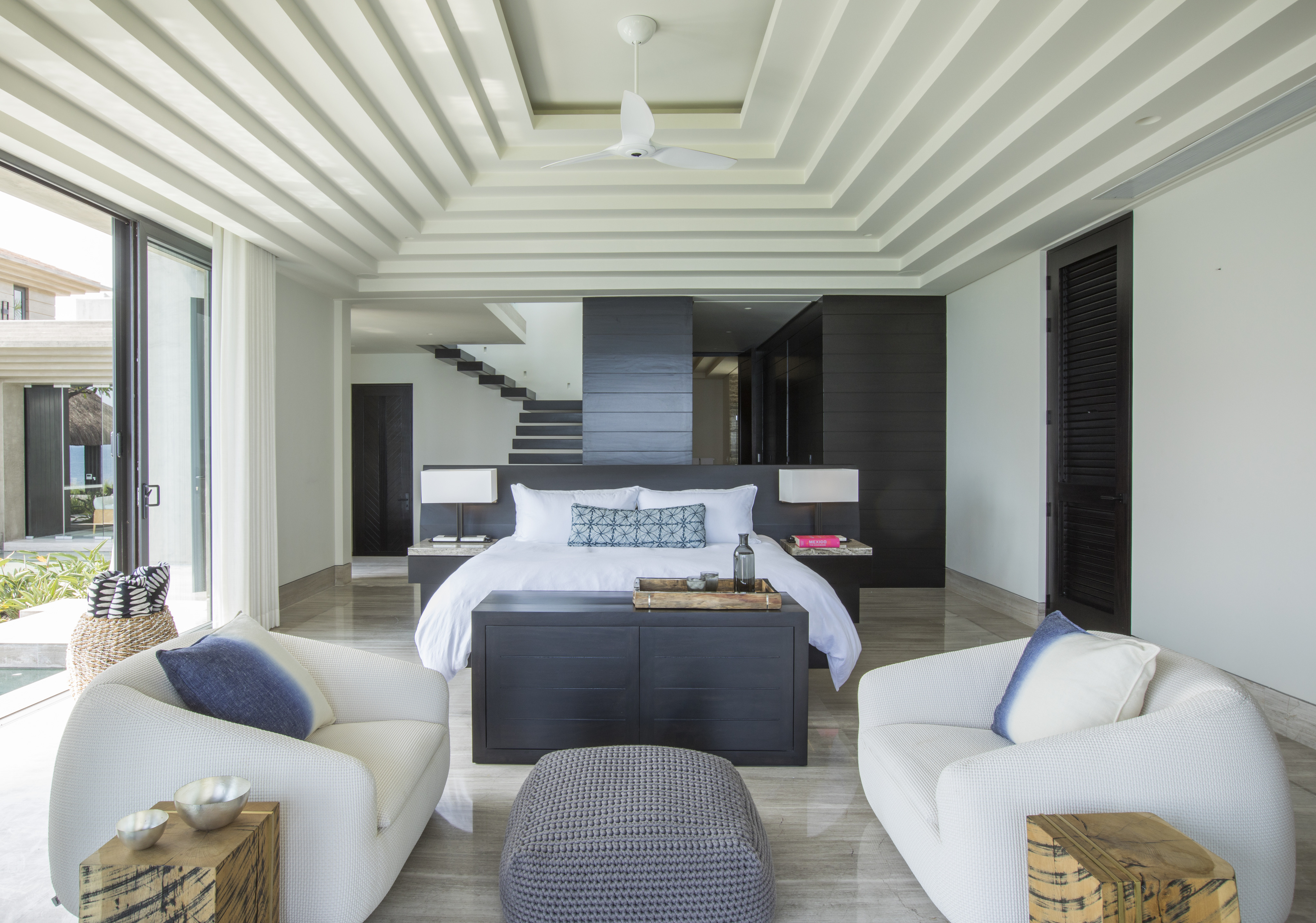 Modern bedroom at La Cascada with a large bed, two white armchairs, a small ottoman, and sliding glass doors opening to an outdoor area. The room features minimalistic decor with a neutral color palette.