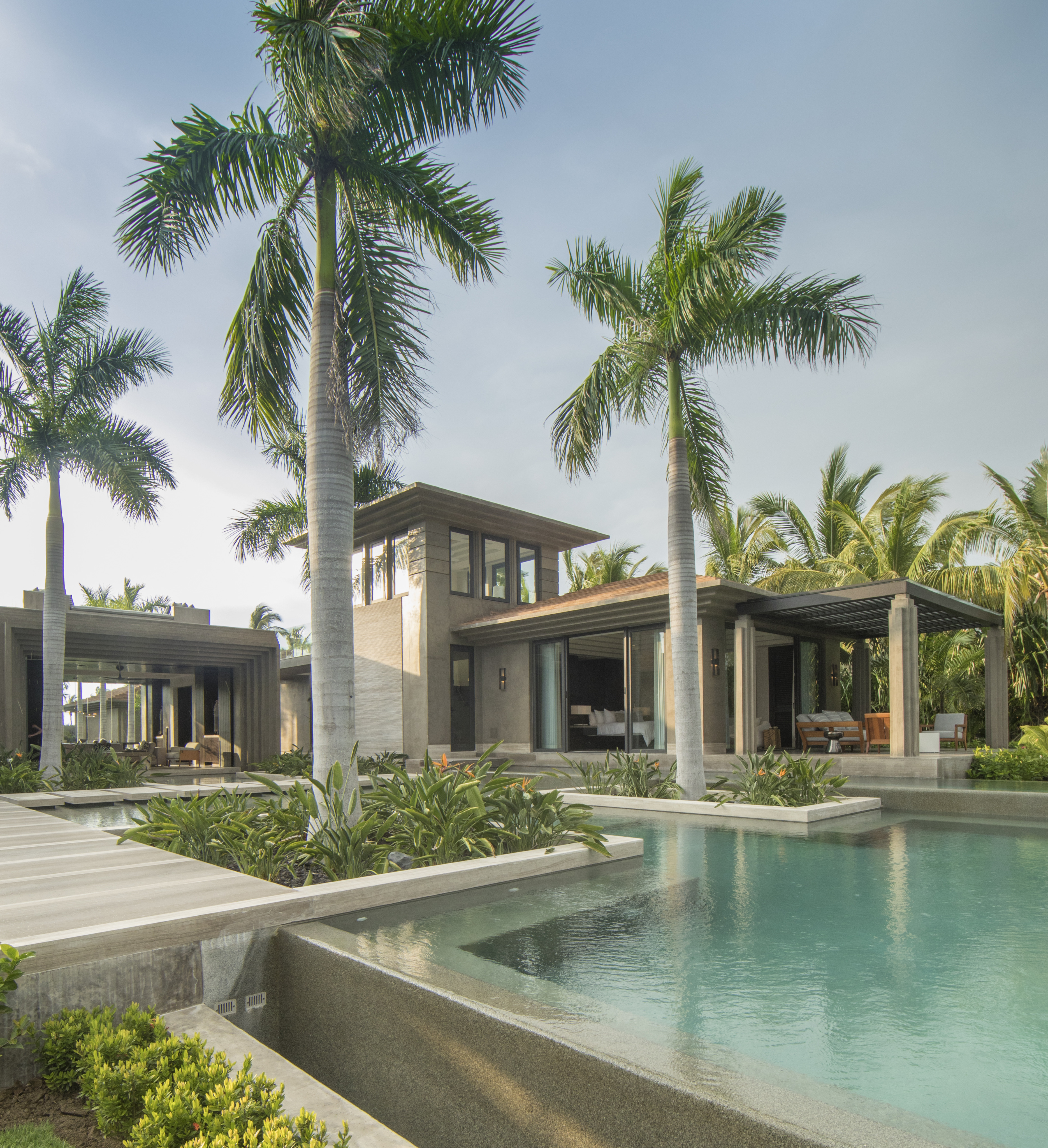 Modern luxury house, La Cascada, surrounded by palm trees, with expansive glass windows, stone and wood exterior, and a large pool in the foreground.