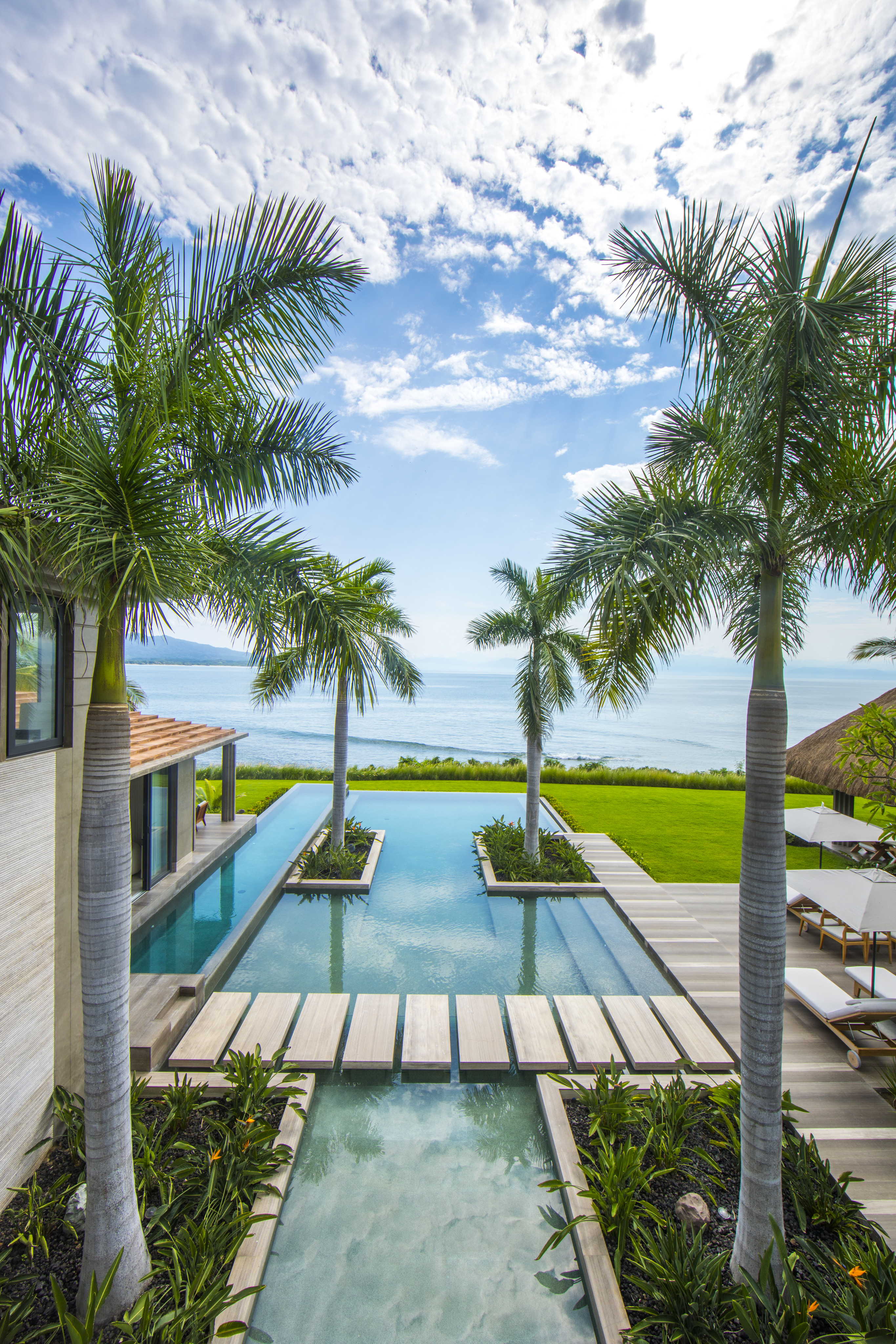 La Cascada is a luxury beachfront property with a pathway over water, surrounded by palm trees and overlooking an ocean beneath a partially cloudy sky.