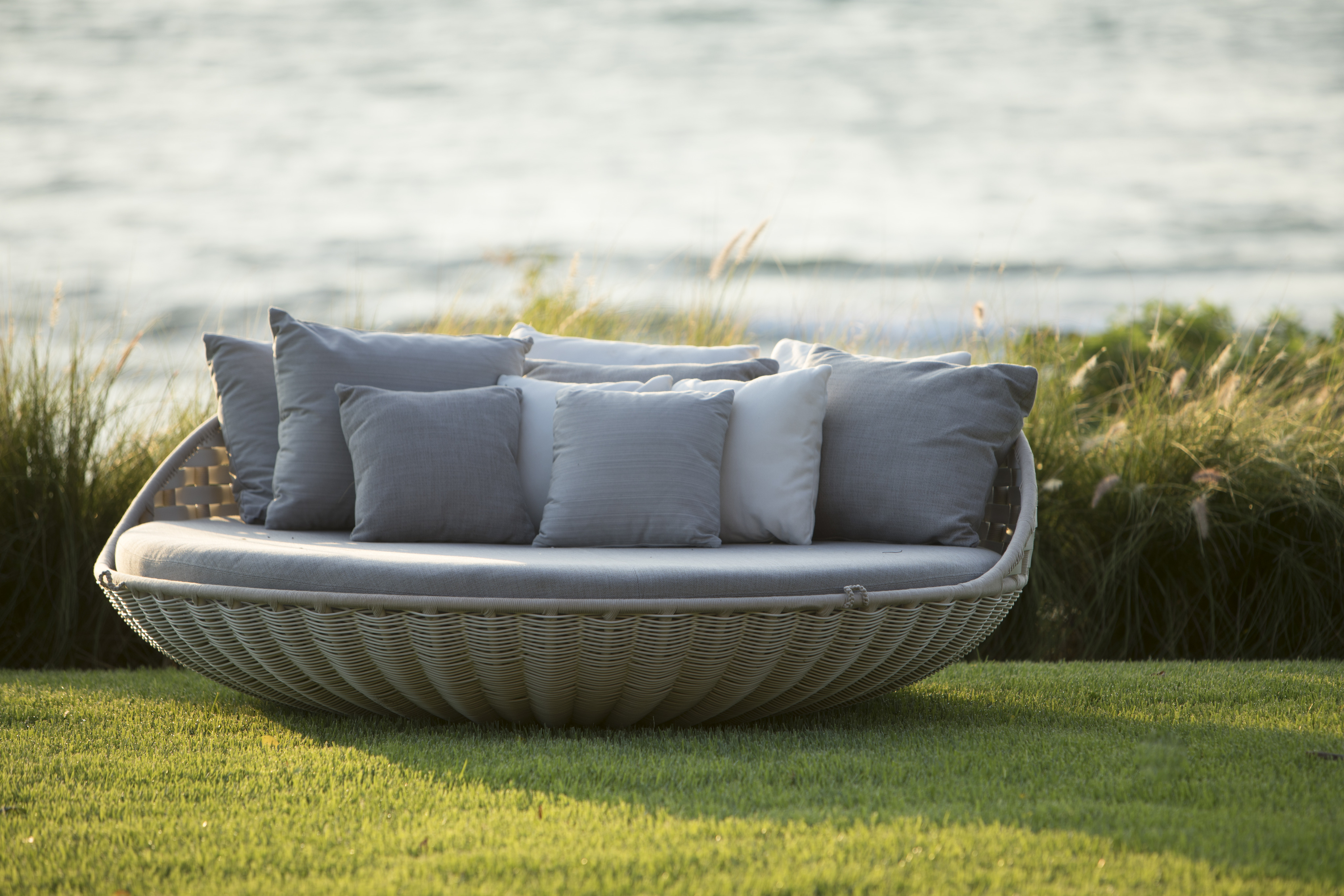 A round wicker outdoor lounge chair with several gray and white cushions, reminiscent of the serene ambiance of La Cascada, is placed on green grass with a backdrop of ocean waves and tall grass.