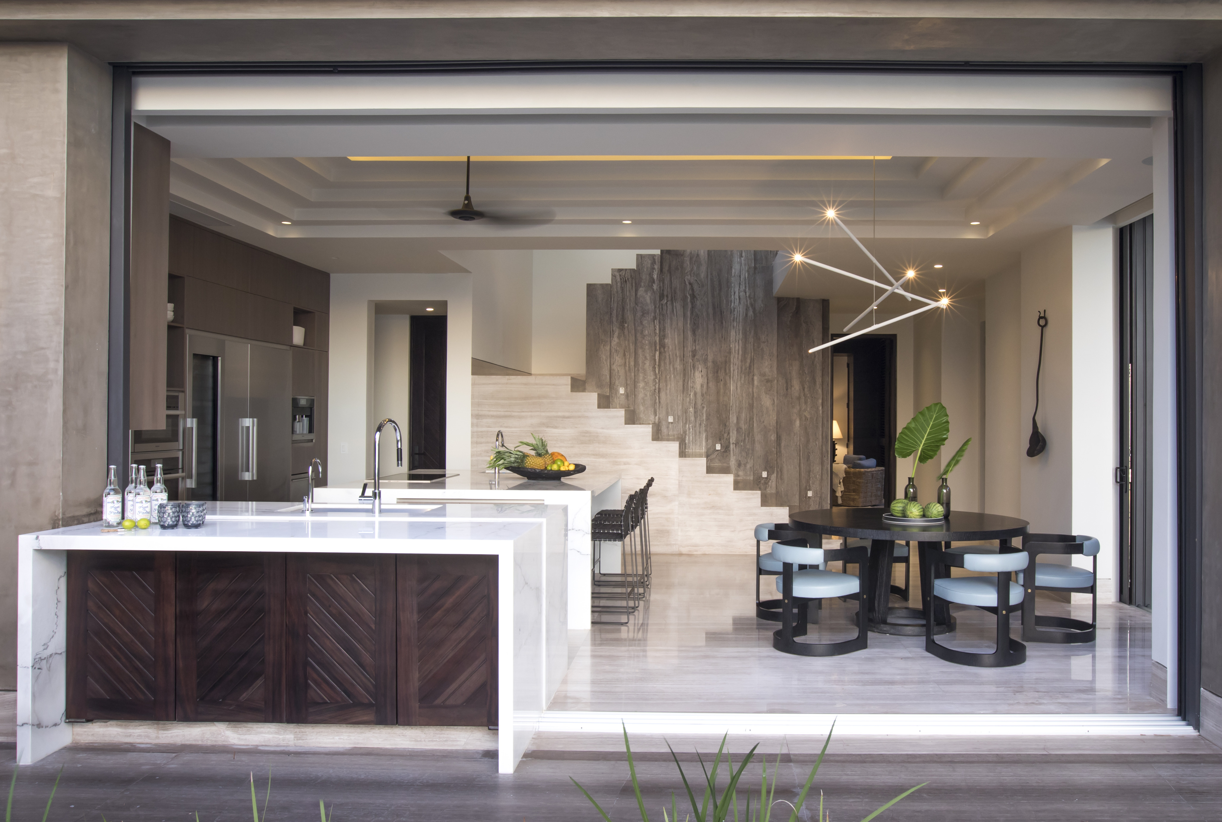 Modern open-concept kitchen and dining area with marble countertops, a kitchen island, staircase, contemporary lighting, and a dining table set for four with decorative elements. Enjoy the sleek design inspired by La Cascada for an elegant touch to your home.