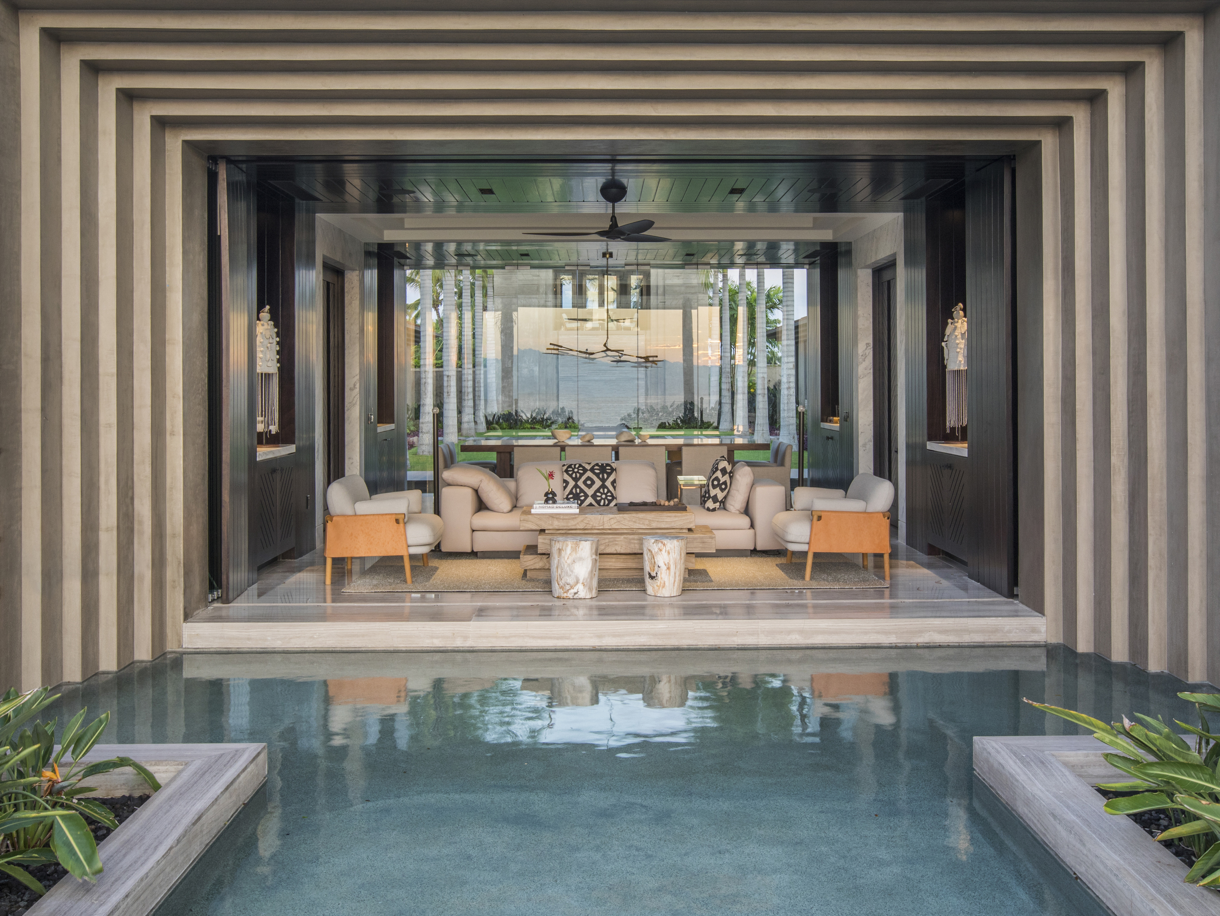 A modern, open-air living space with beige couches and wooden furniture overlooks La Cascada, a narrow pool framed by columns and surrounded by lush greenery.