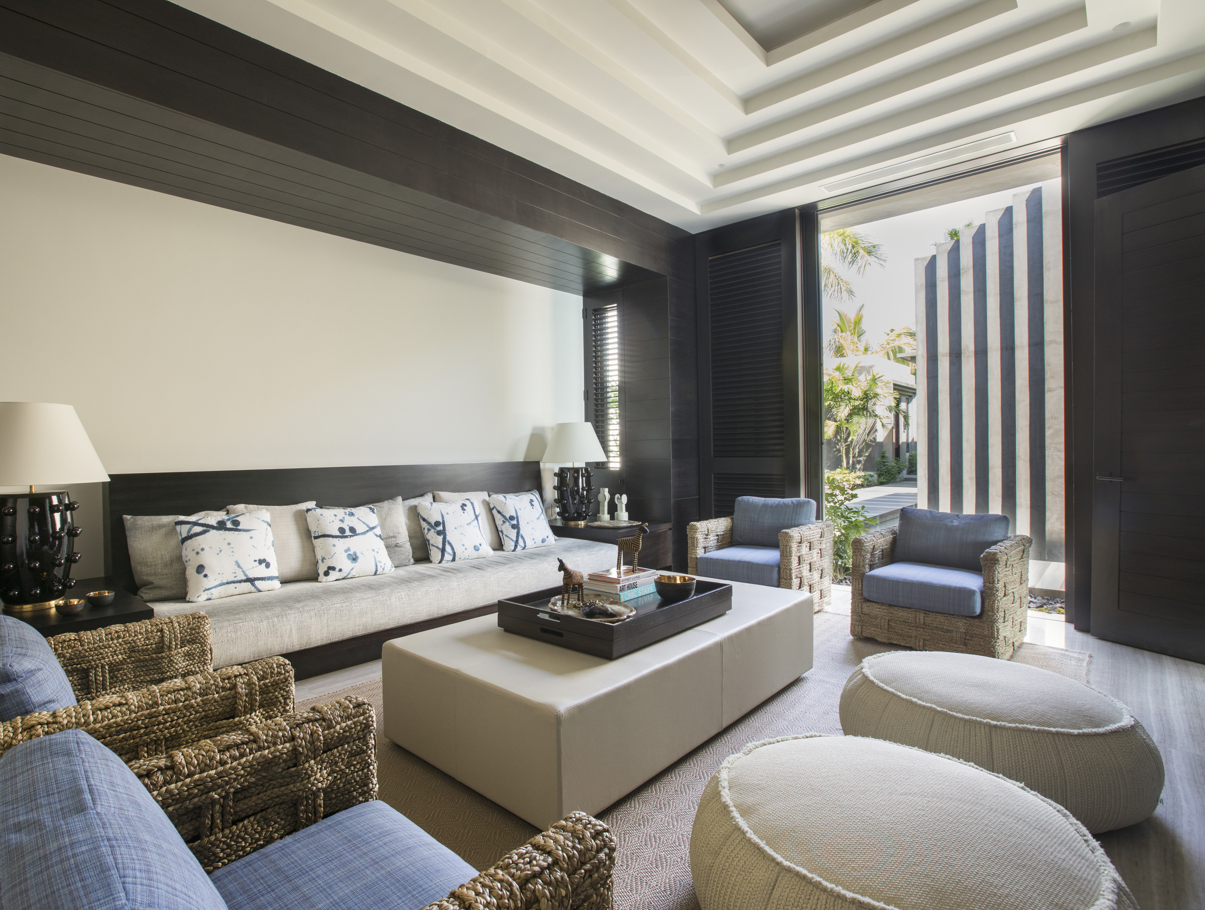 Modern living room with cream-colored furniture, light blue accents, and wicker chairs. Open sliding doors reveal a lush garden outside. Two large ottomans and a tray on the central ottoman table enhance this La Cascada-inspired space.