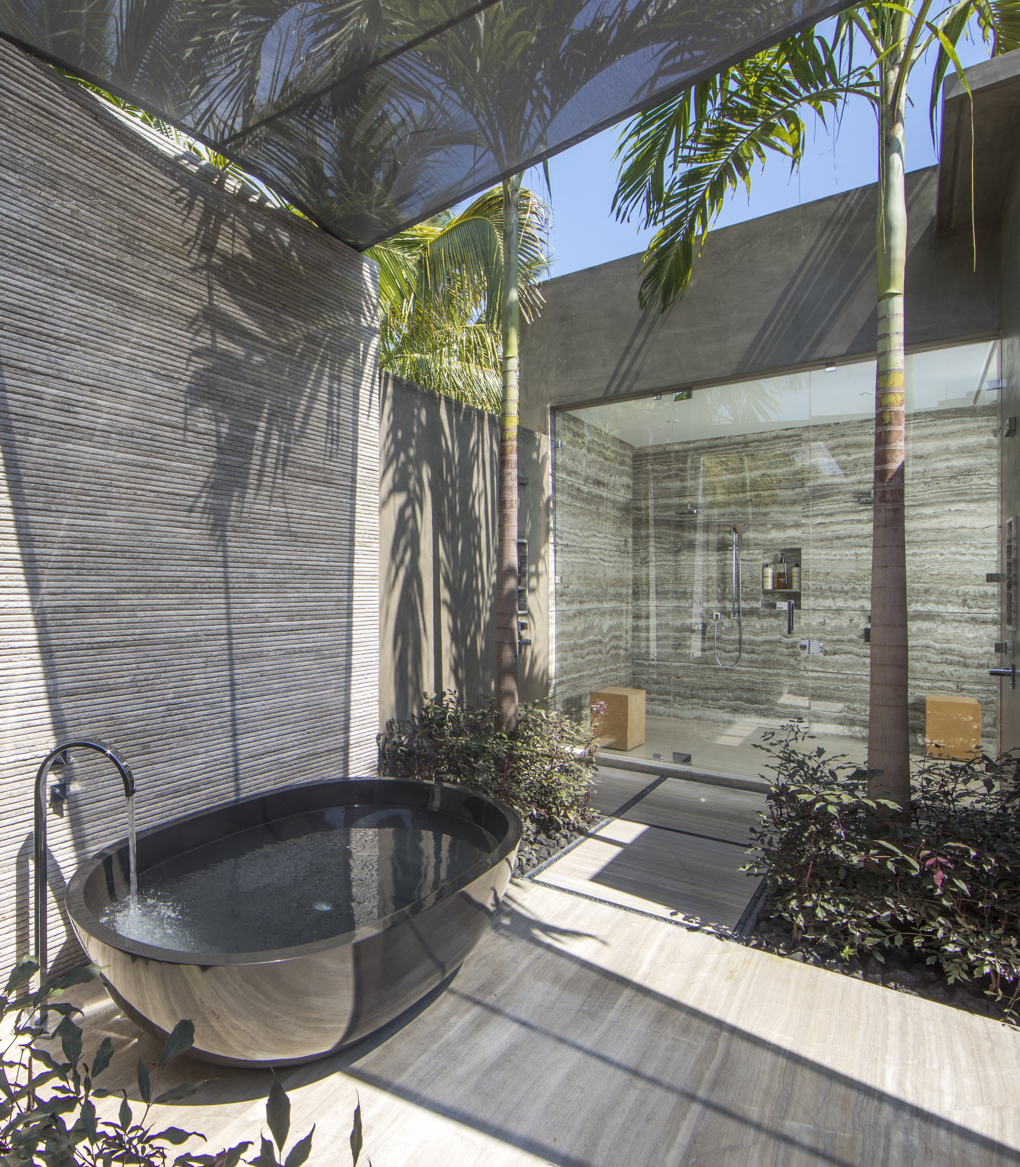 Modern outdoor bathroom with a freestanding black tub, surrounded by plants. Adjacent to the tub is an enclosed shower area with glass walls and contemporary fixtures. Palm trees provide shade overhead, creating a serene escape reminiscent of La Cascada's tranquil ambiance.