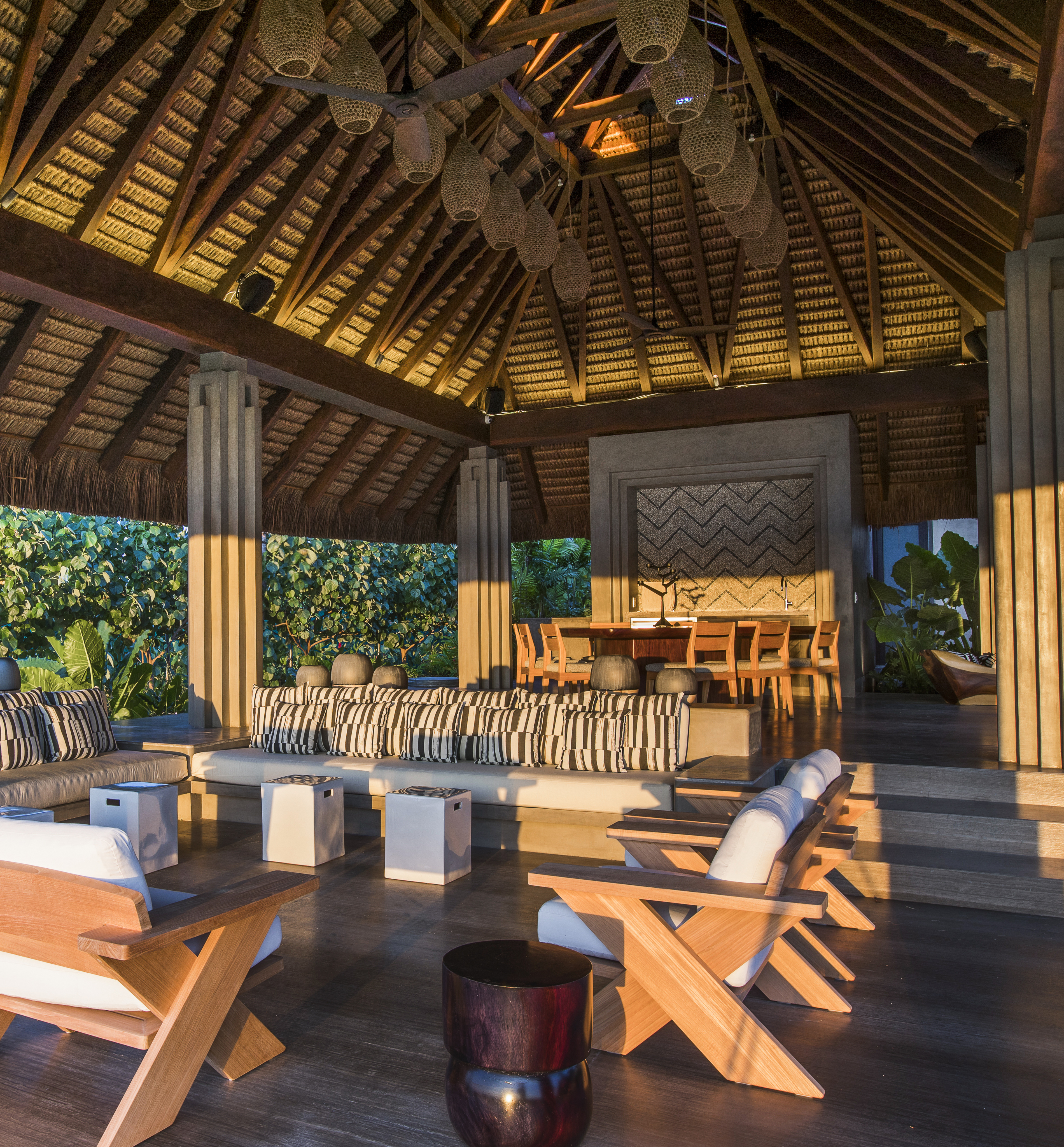 La Cascada features an open-air lounge with wooden furniture, striped cushions, a high thatched roof, and a dining area in the background, all surrounded by dense greenery.