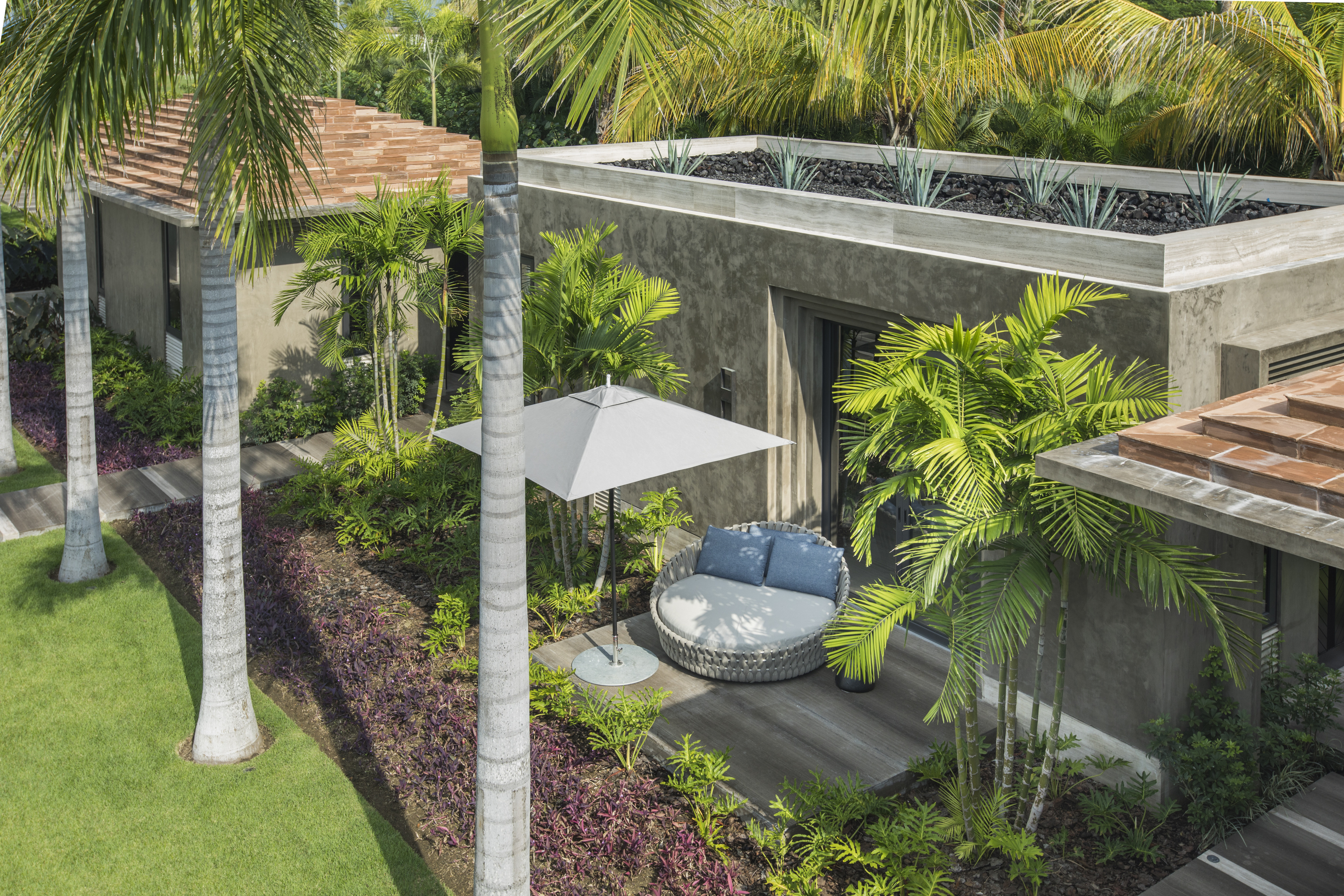 Modern outdoor seating area with a circular couch and umbrella amid lush greenery and palm trees, adjacent to the contemporary La Cascada building with a rooftop garden.