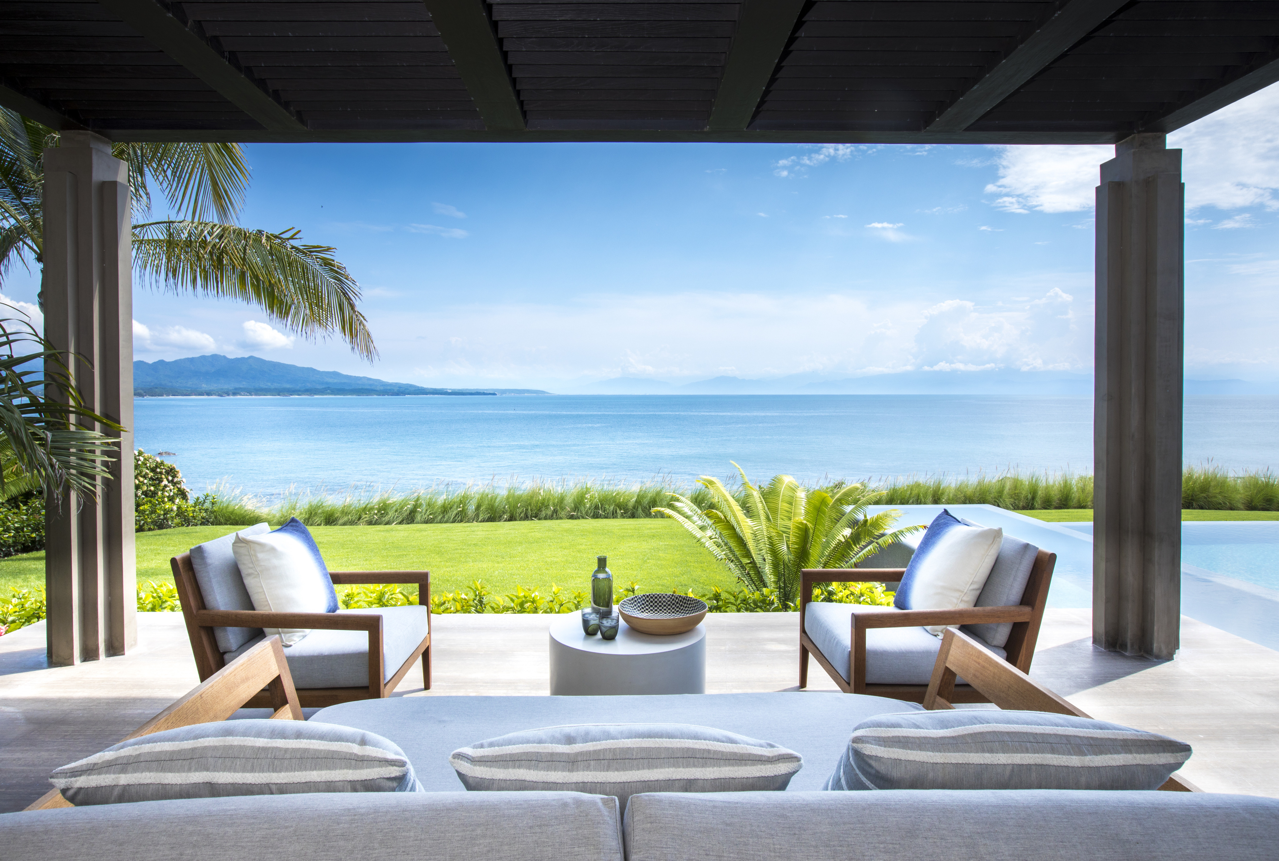 Outdoor seating area at La Cascada with a panoramic ocean view. Two chairs and a sofa surround a small table with a bottle and two glasses, framed by palm trees and lush greenery.