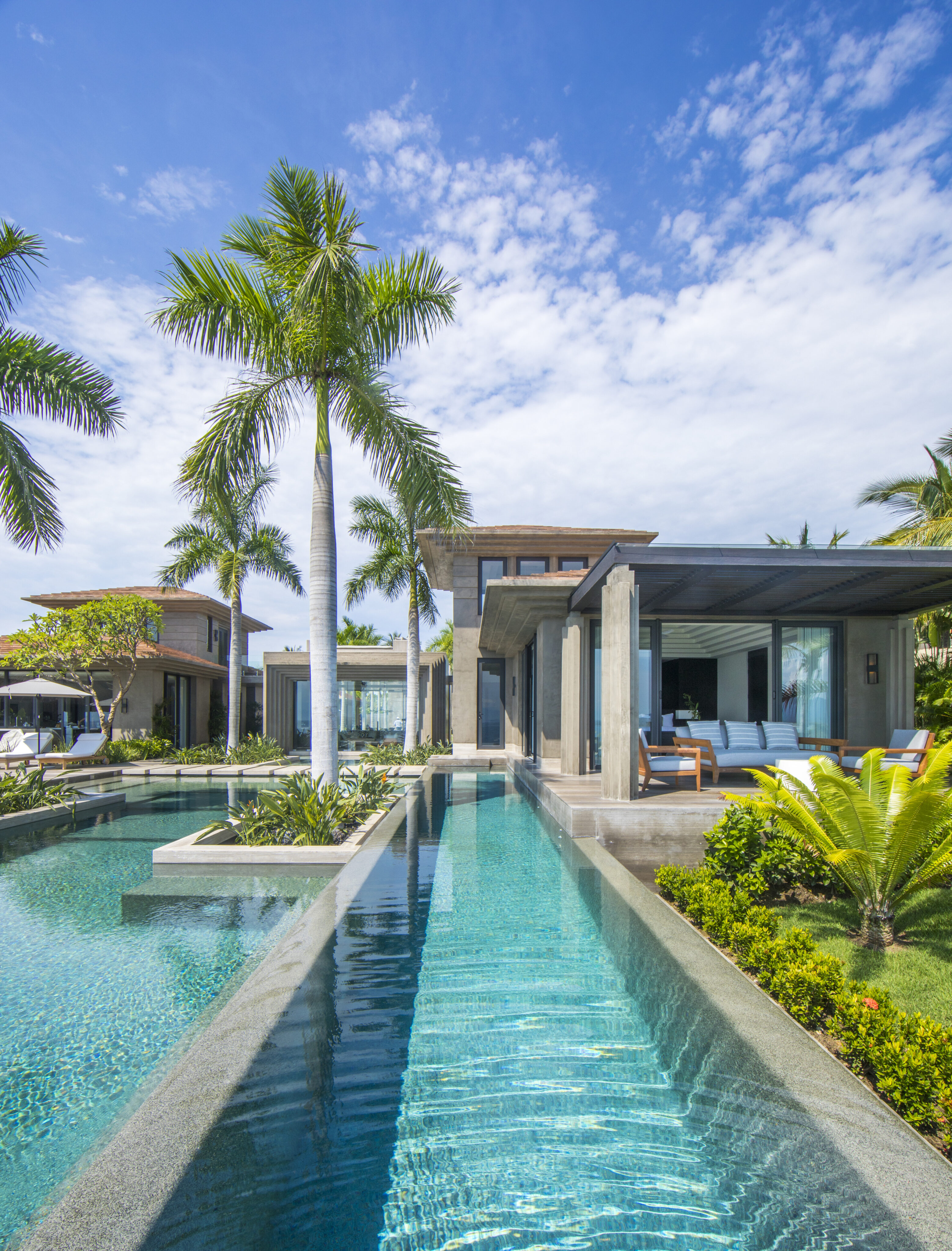 A luxurious modern villa, La Cascada, with a swimming pool surrounded by palm trees, set against a clear blue sky. The villa features large glass windows and a spacious open living area with outdoor seating.