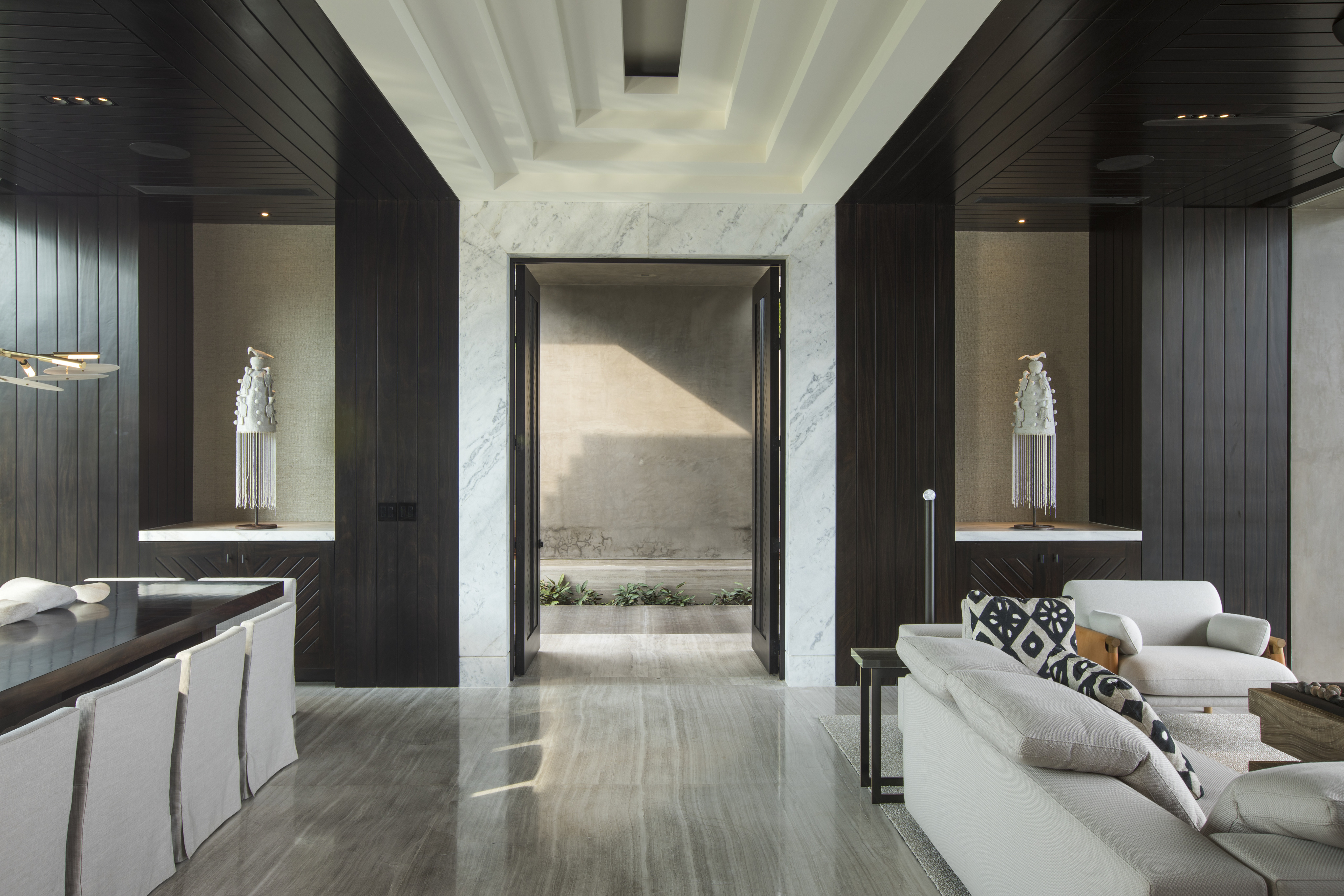 A modern living room, known as La Cascada, features light-colored marble floors, dark wood paneling, and white furniture. A central doorway opens to a small, sunlit courtyard with minimalistic decor.