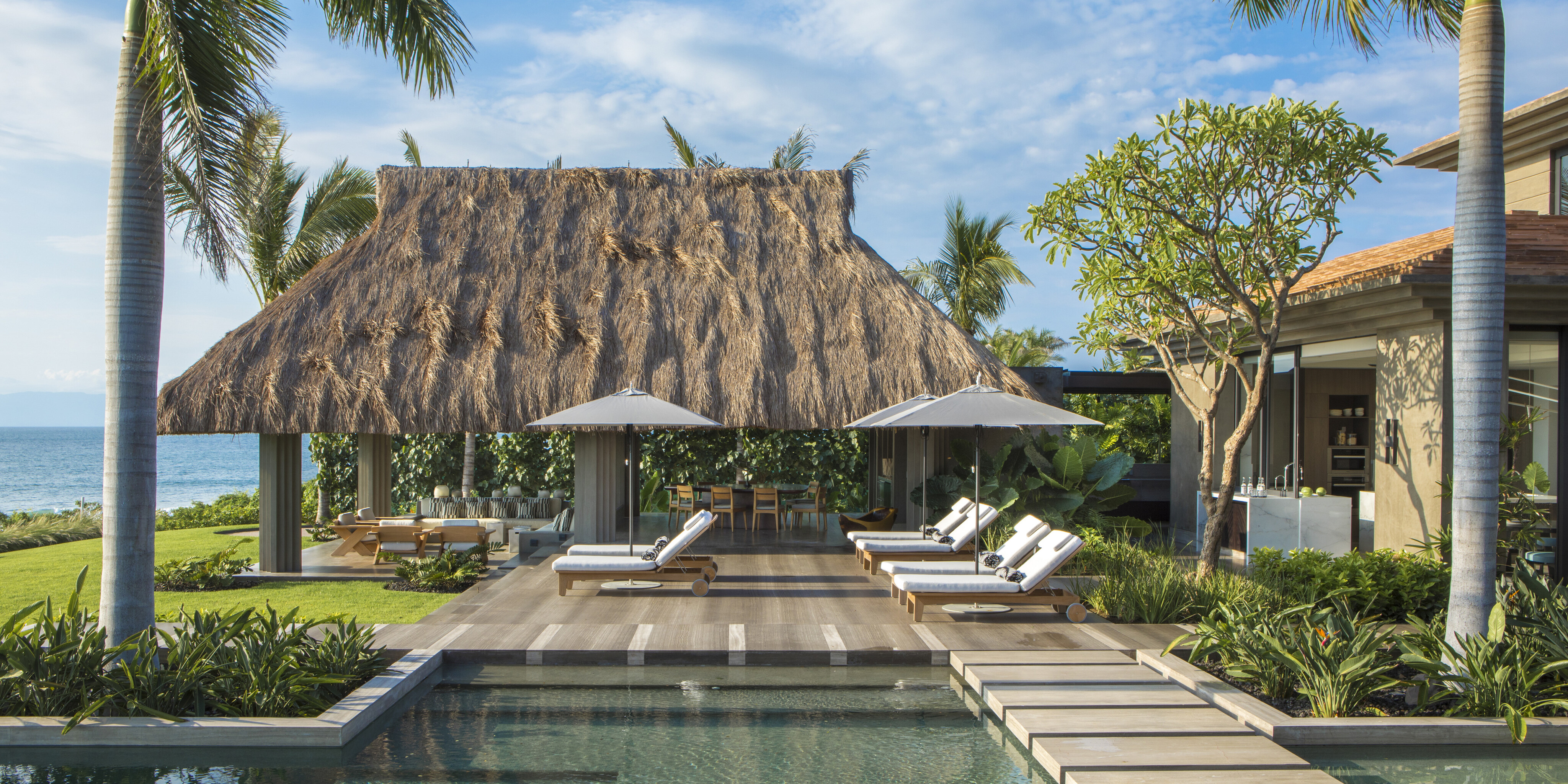 A luxurious outdoor area with a thatched-roof gazebo, lounge chairs, sun umbrellas, a swimming pool, and tropical landscaping, set against a backdrop of the ocean and a bright blue sky at La Cascada.