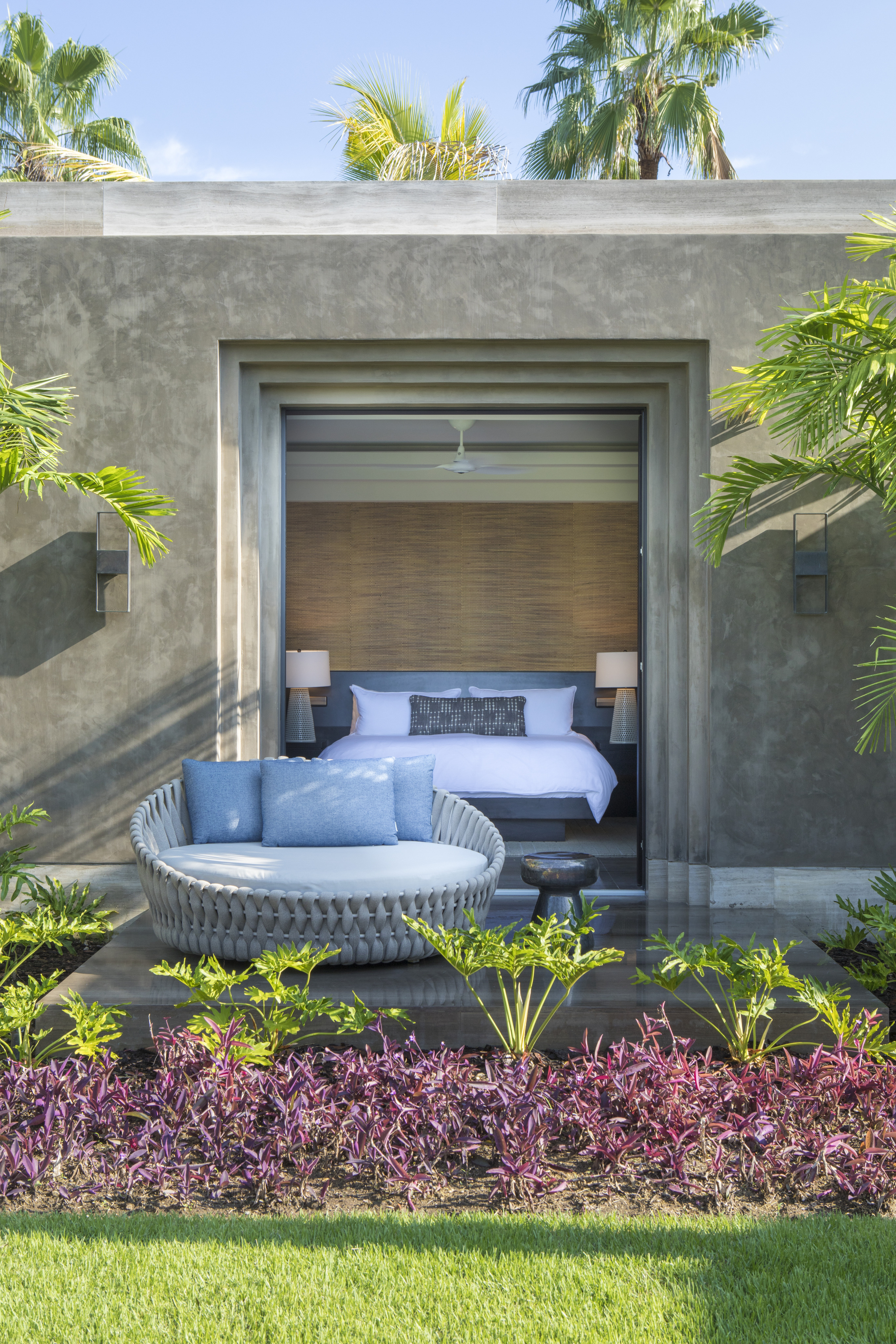 A modern bedroom with open sliding doors leads to an outdoor seating area, featuring a circular sofa by La Cascada. The space is surrounded by tropical plants and greenery.