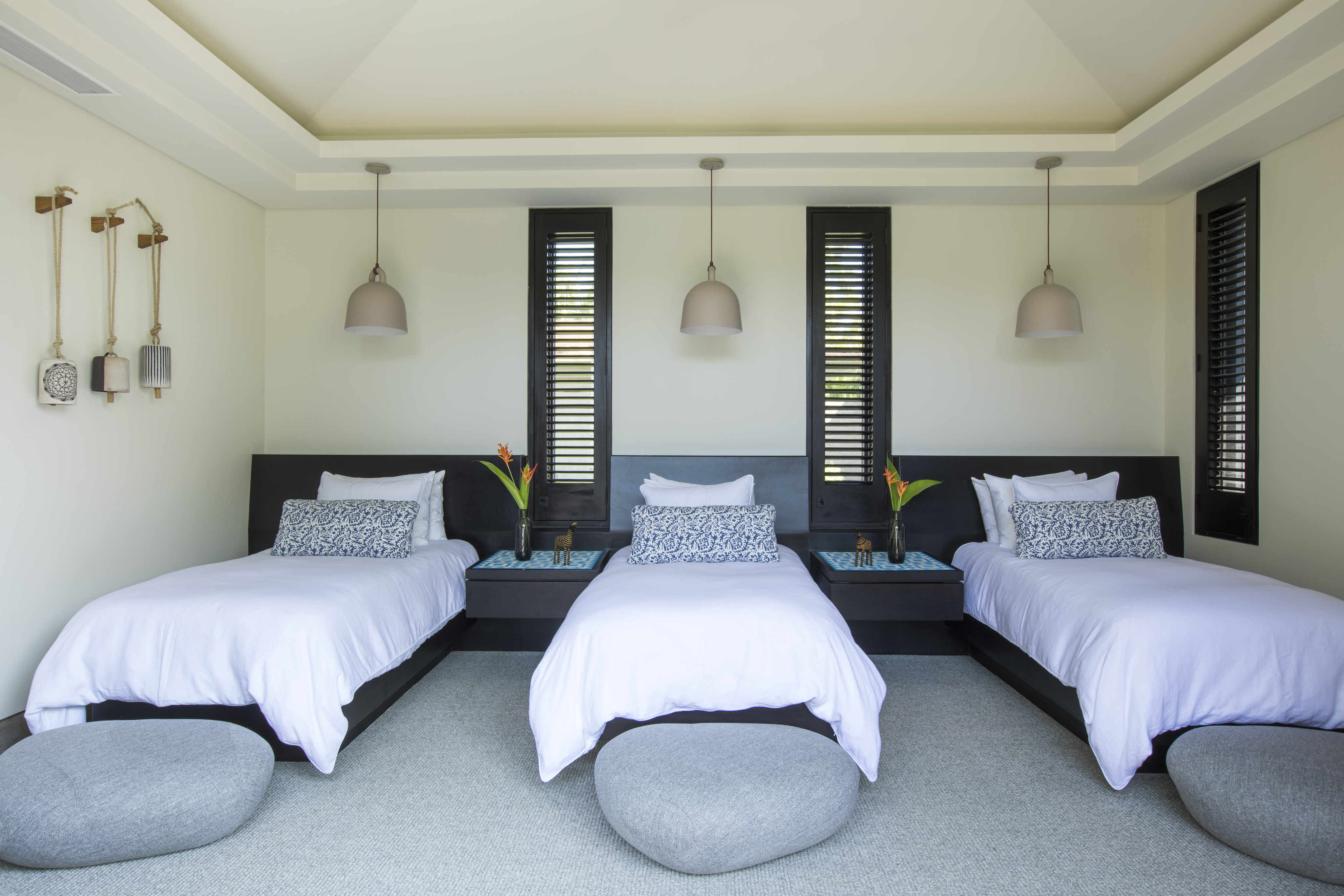 A minimalist bedroom inspired by La Cascada features three single beds aligned in a row, each with white bedding and gray headboards. Round stools are placed in front of each bed. Two windows with shutters adorn the back wall, allowing natural light to flood the serene space.