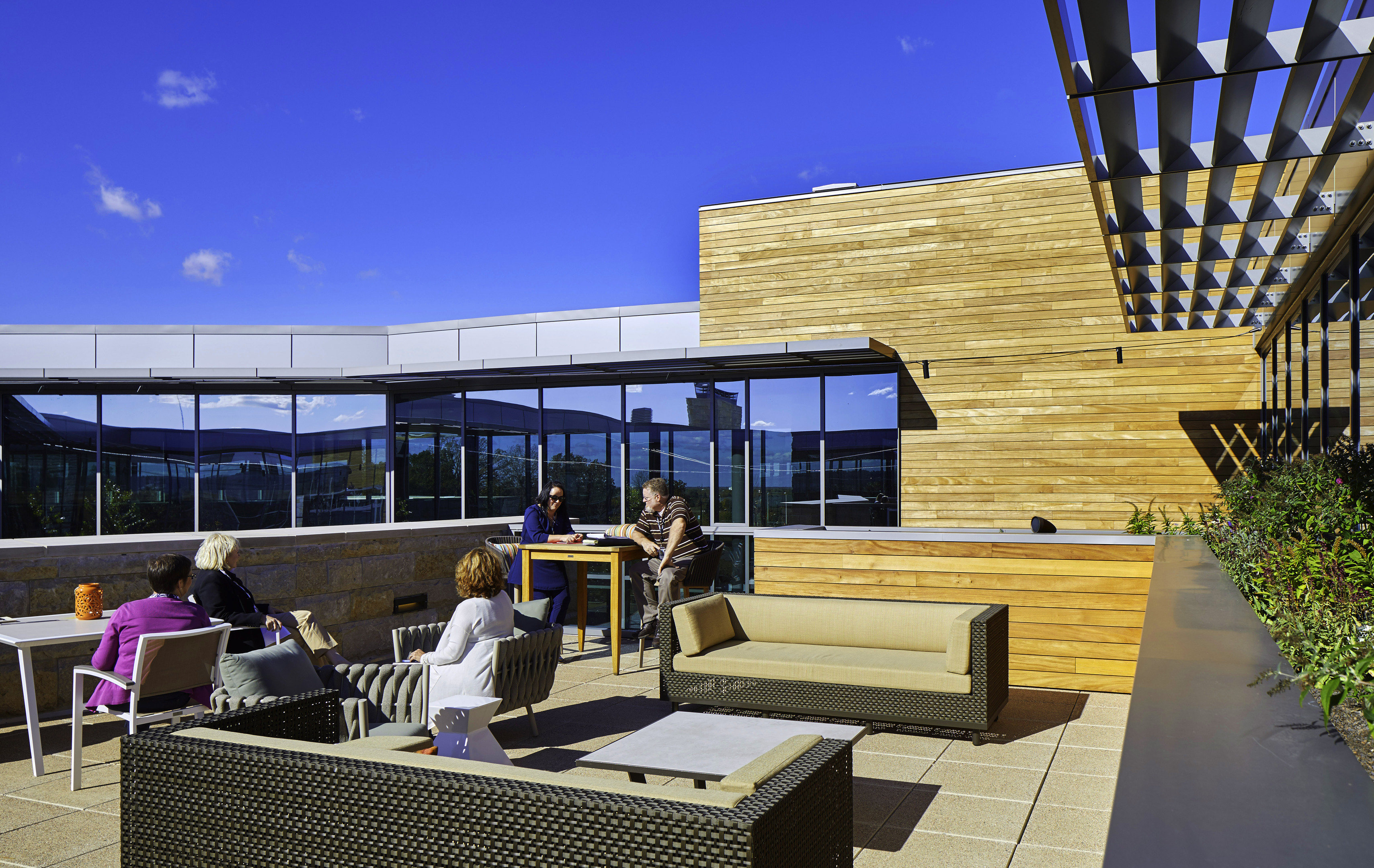 Four people sit and converse on the rooftop terrace of a modern research center with wooden walls and large windows. The area is furnished with comfortable outdoor seating and tables under a clear blue sky.